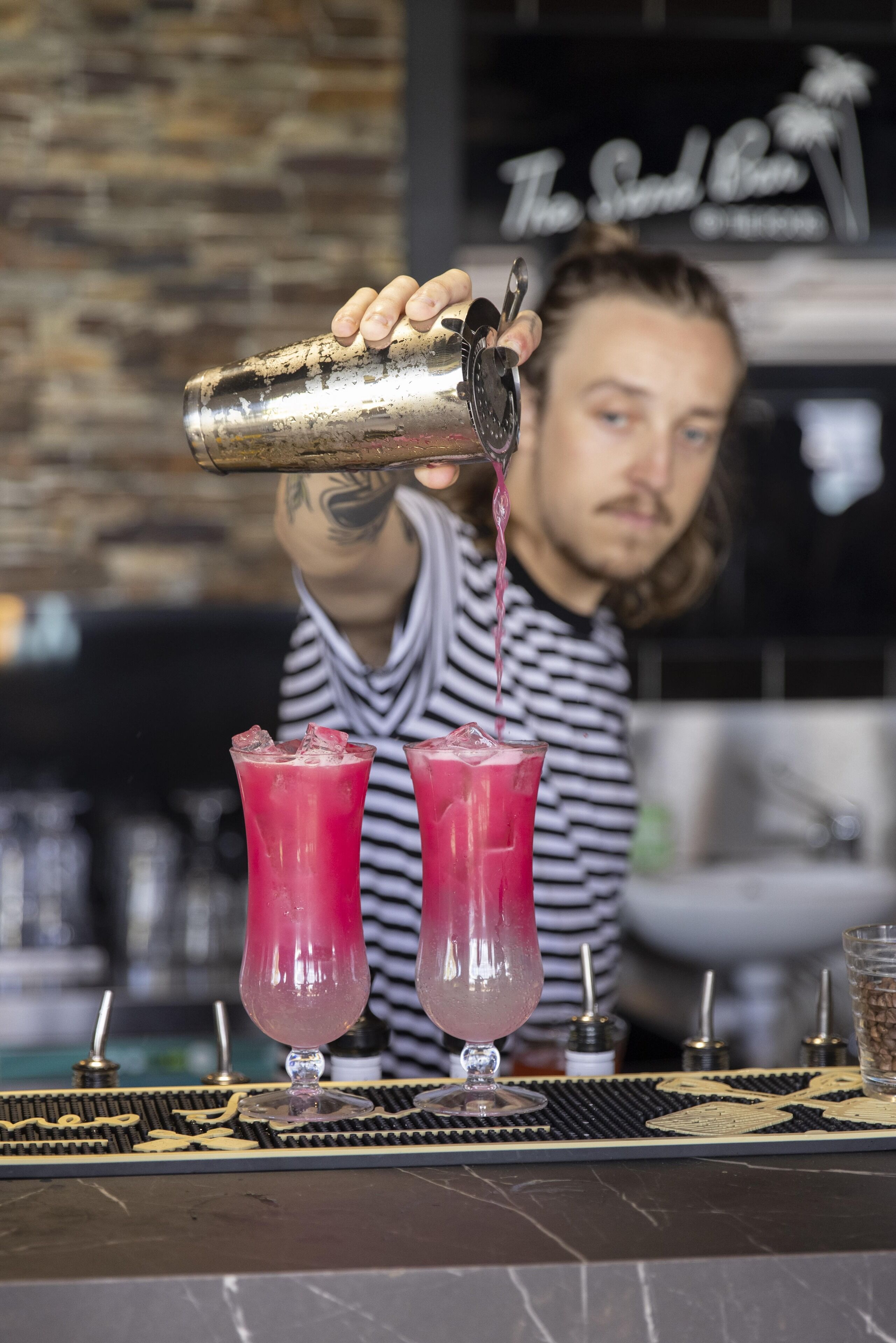A bartender pouring a pink drink into two glasses on a bar counter.