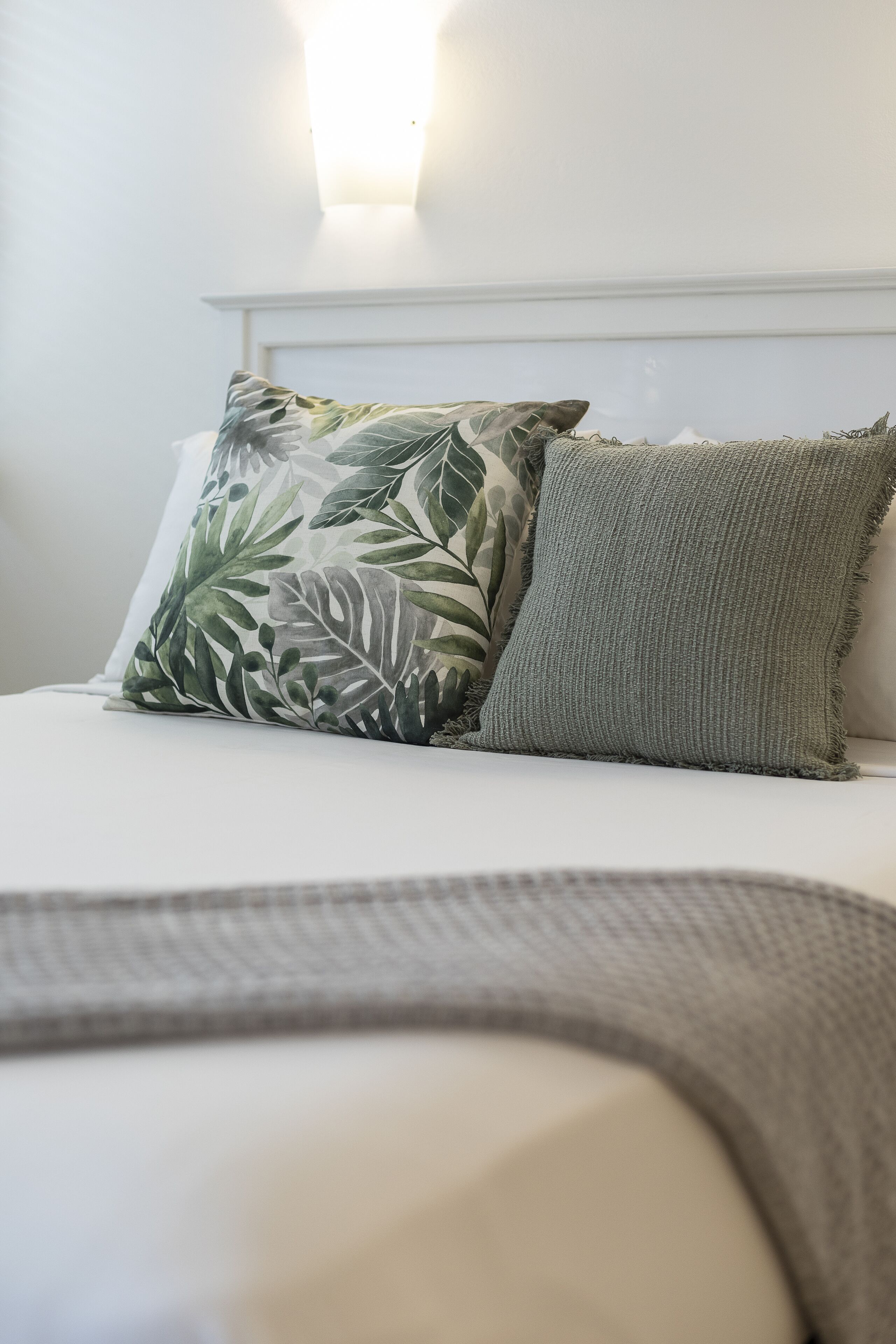 A bed with two pillows, one with a leaf pattern and one plain, in a room with a white headboard and a wall-mounted light fixture.