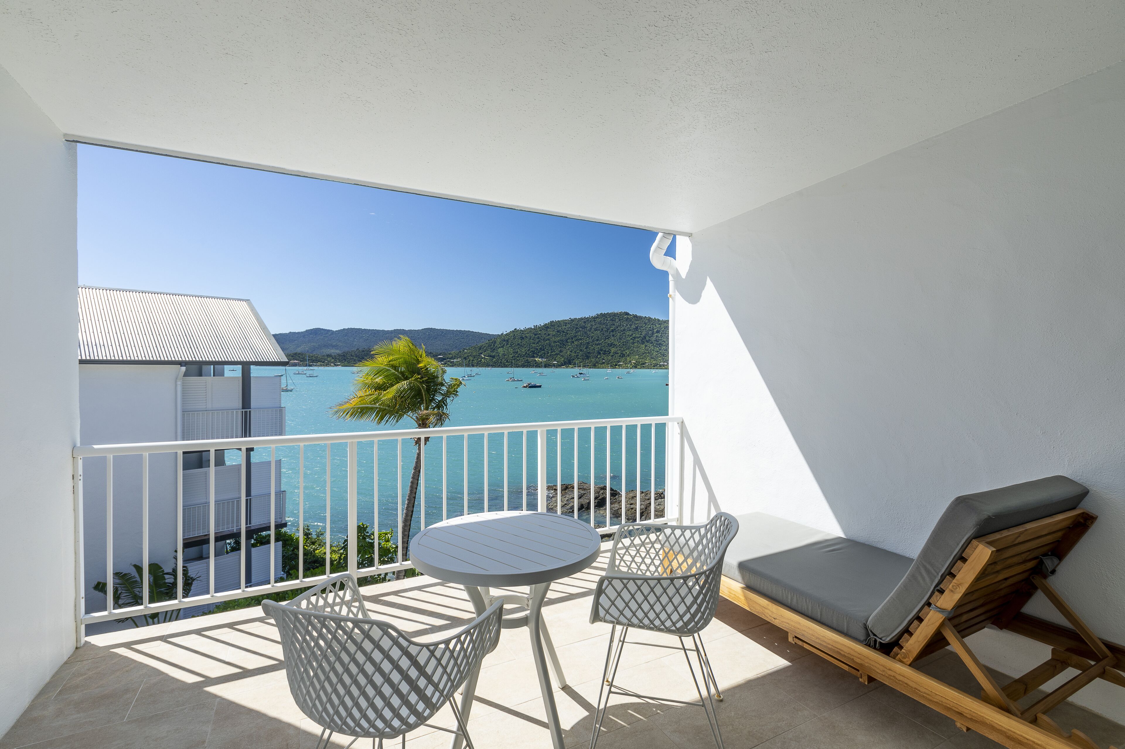 A balcony with a view of the sea, a table, two chairs, and a wooden lounge chair.
