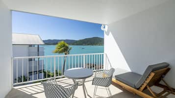 A balcony with a view of the sea, a table, two chairs, and a wooden lounge chair.