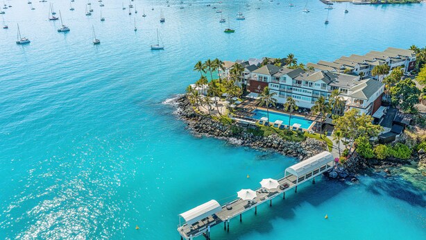 Vista aérea de un hotel frente al mar con piscina, muelle y varios barcos en el agua.