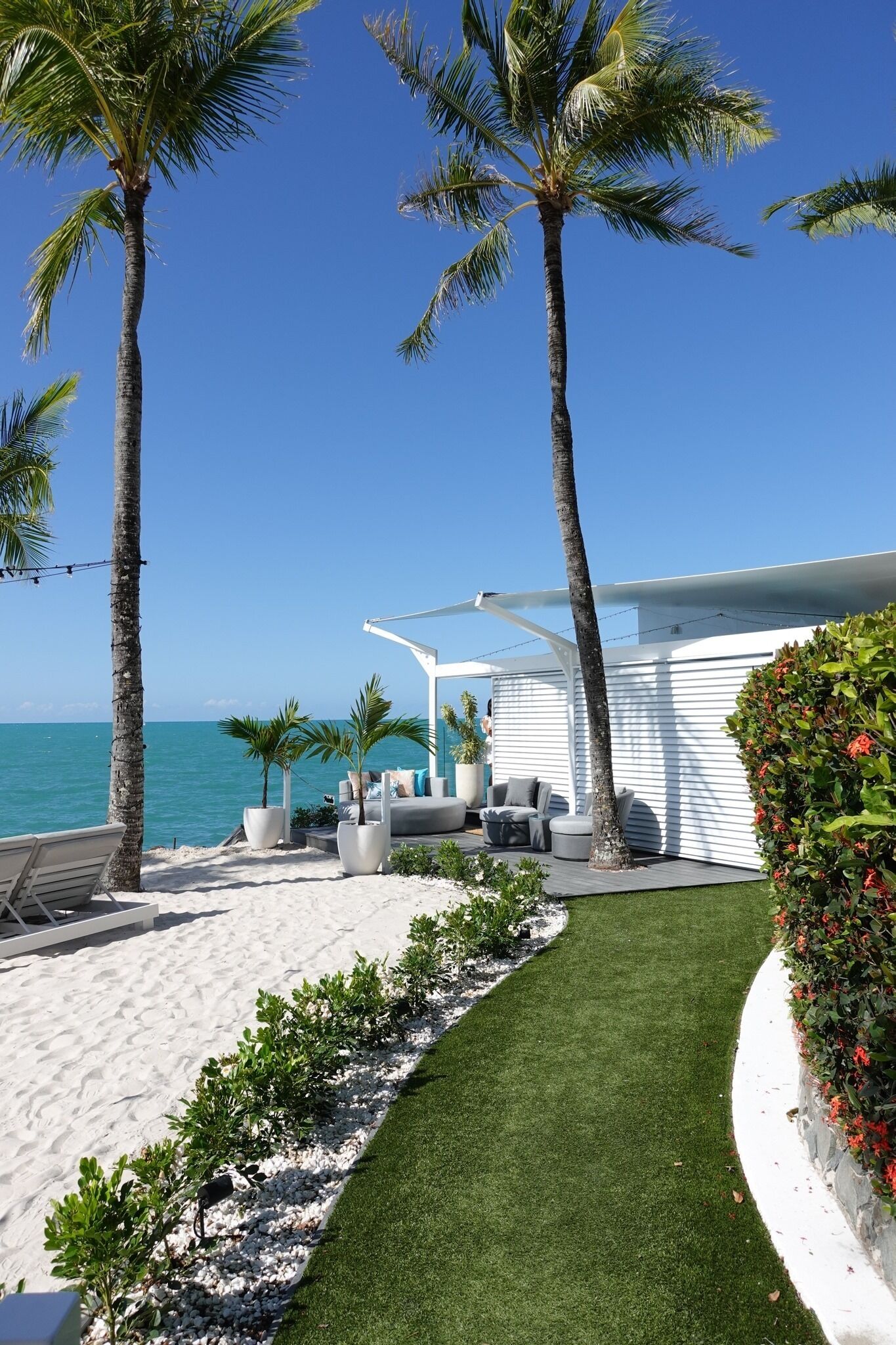 A beachside patio with a white pergola, outdoor seating, and palm trees.