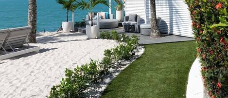 Un patio frente a la playa con una pérgola blanca, mobiliario exterior y palmeras.