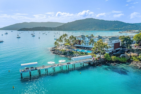 A coastal hotel with a pier, a pool, and a view of the sea and hills.