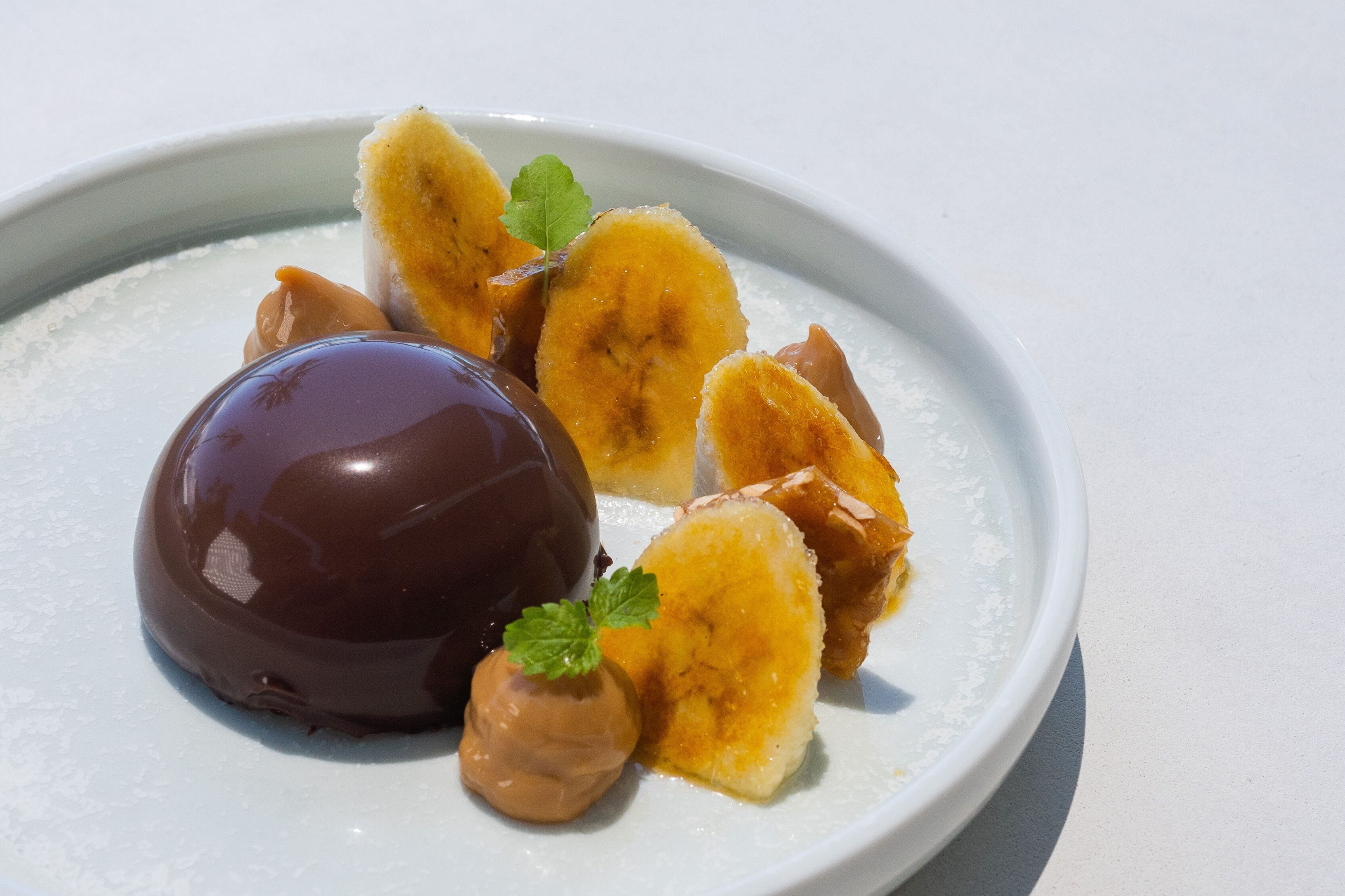 A dessert plate with a chocolate sphere, banana slices, and caramel dollops.
