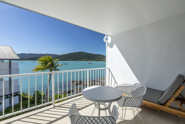 A balcony with a view of the sea, a table, two chairs, and a sun lounger.