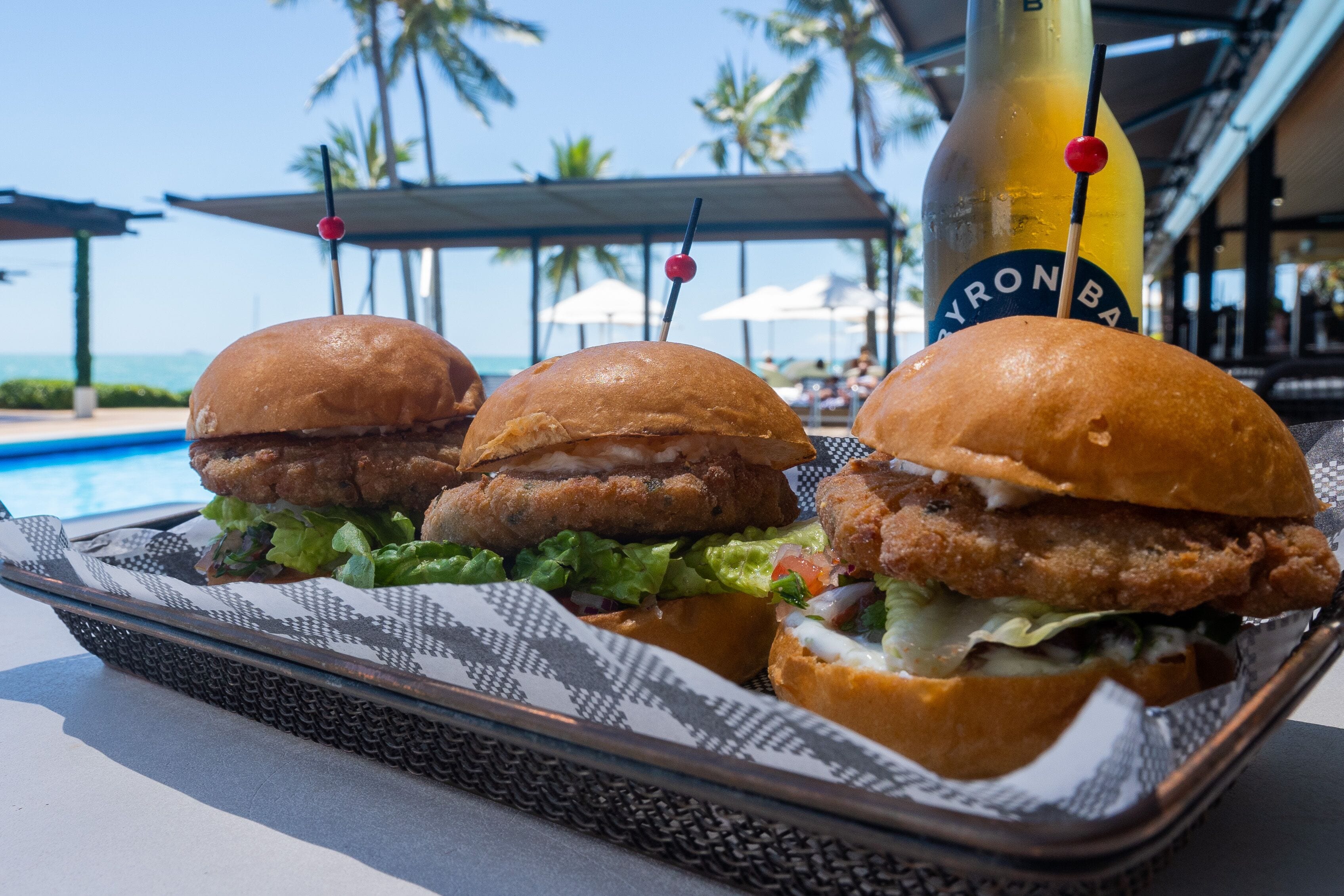 A tray with three burgers and a bottle of beer on a table by a poolside.
