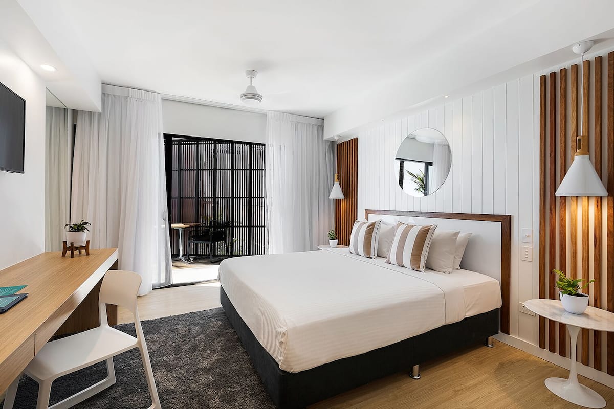 Een moderne hotelkamer met een groot bed, een bureau en een stoel. Er staat een klein tafeltje met een plantje en een spiegel aan de muur.