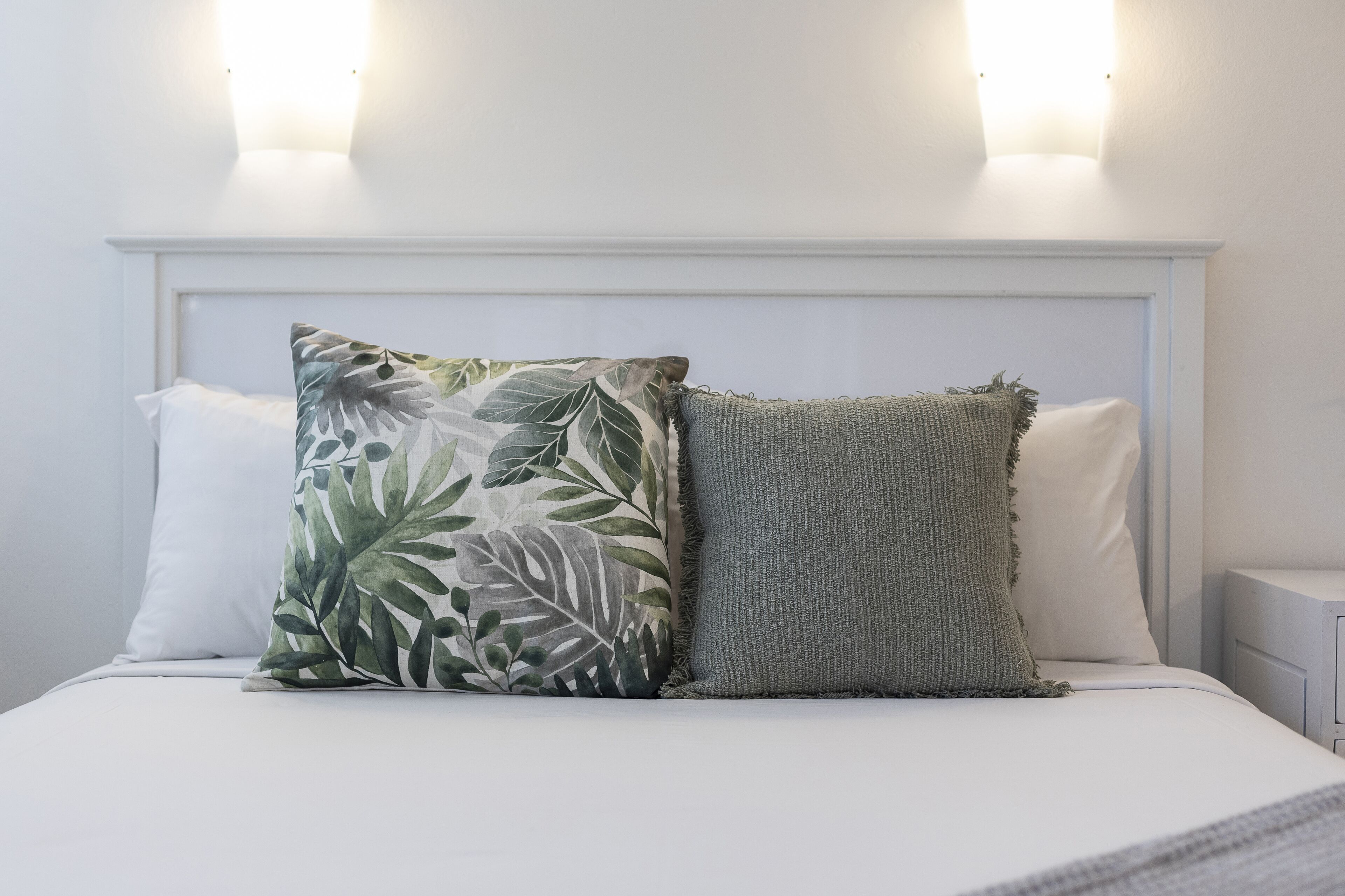 A bed with two pillows, one with a leaf pattern and one plain, in a room with a white headboard and wall-mounted lights.