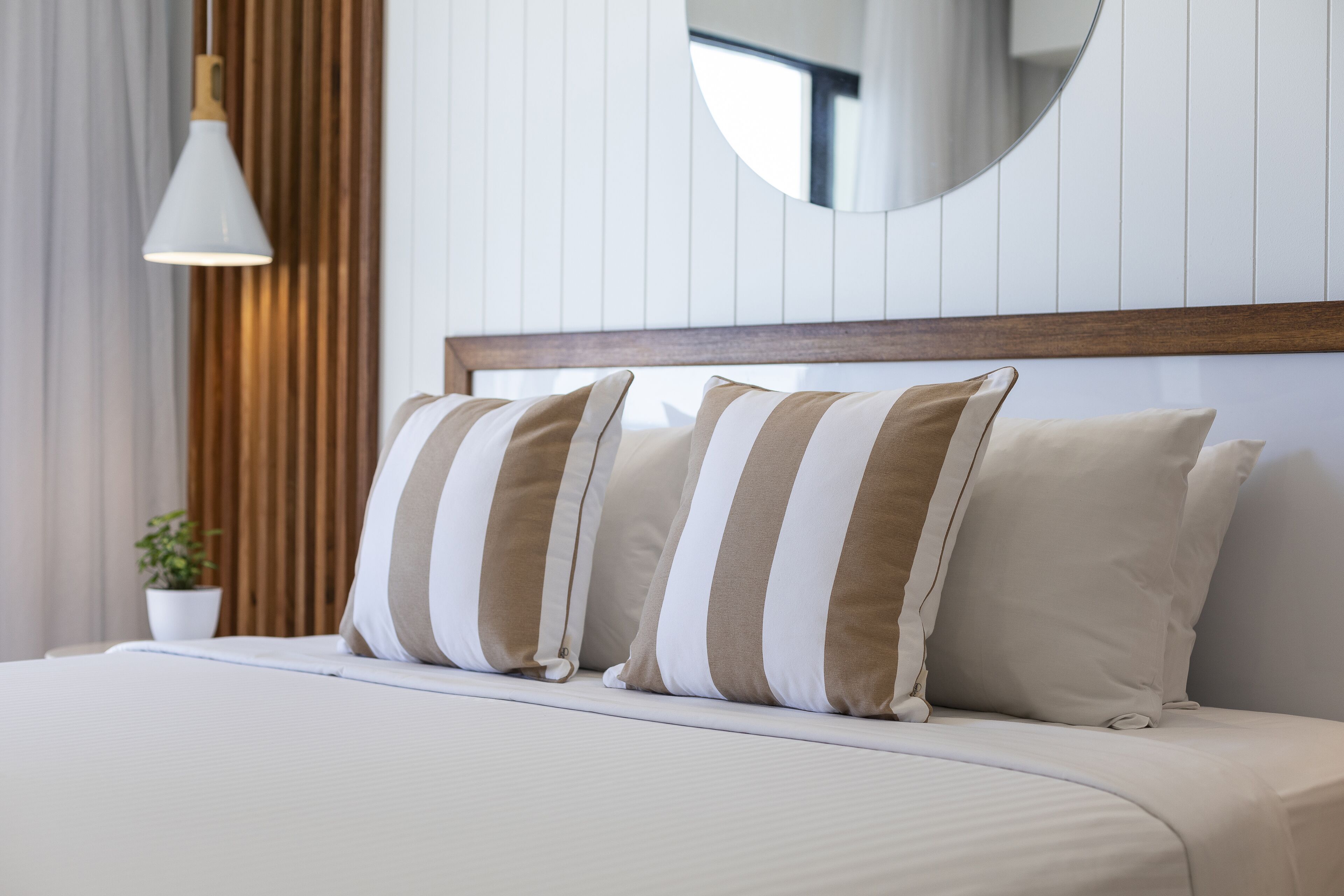 A neatly made bed with striped pillows, a wooden headboard, a hanging light fixture, and a potted plant.