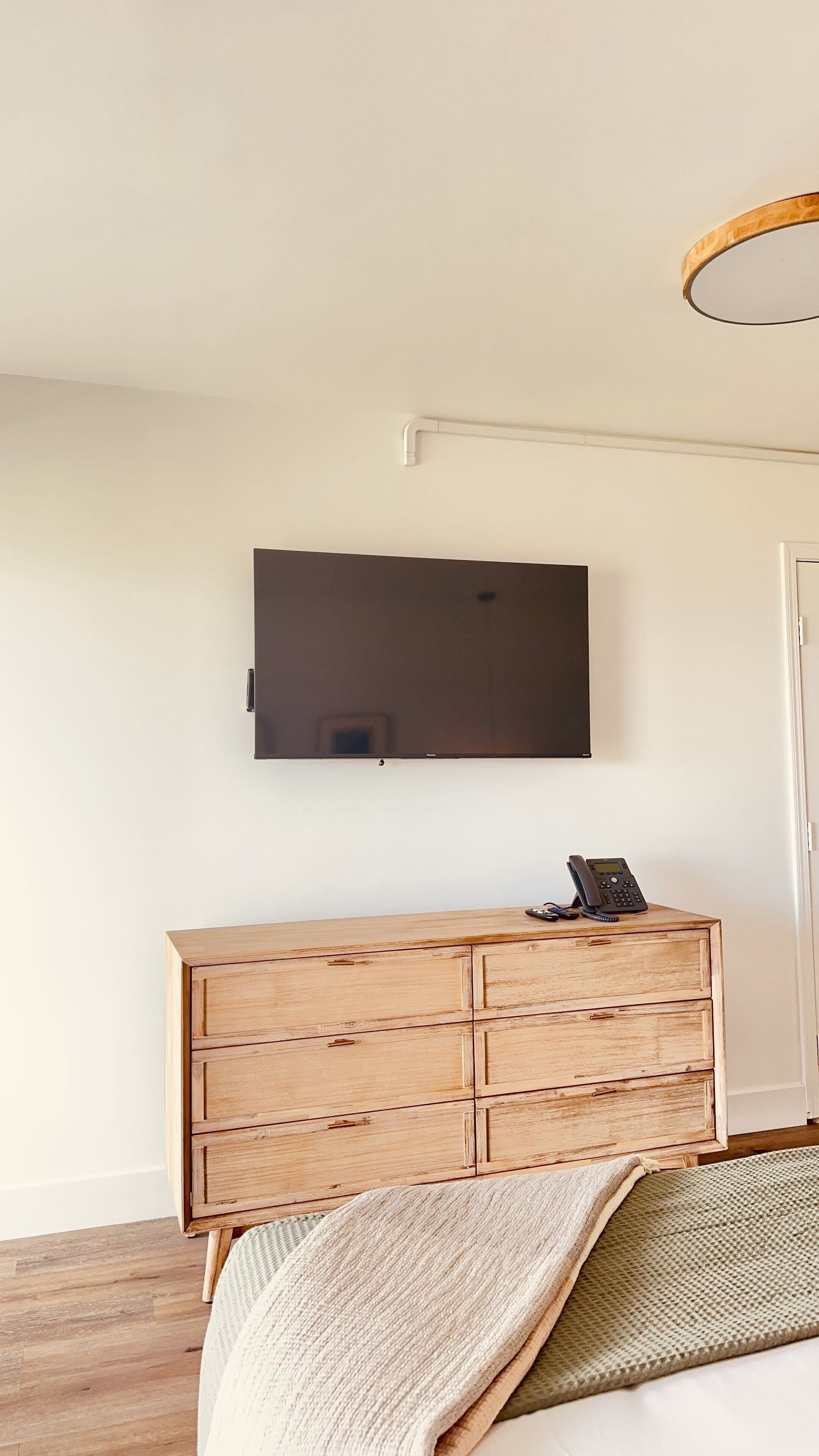 Ein Schlafzimmer mit einem hölzernen Schminktisch, einem an der Wand befestigten Flachbildfernseher und einem Bett mit weißer Decke.