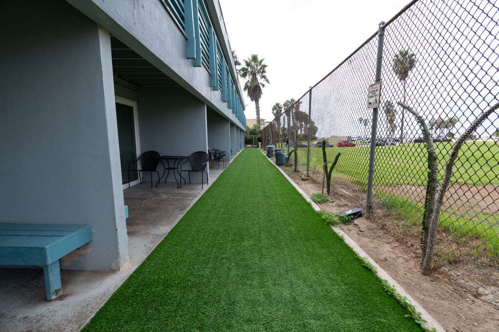 A building with a covered patio area, a green artificial turf path, and a chain-link fence enclosing a sports field.