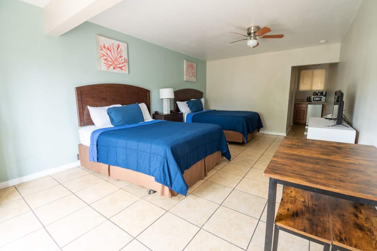 A hotel room with two beds, a wooden table, a ceiling fan, and artwork on the walls.