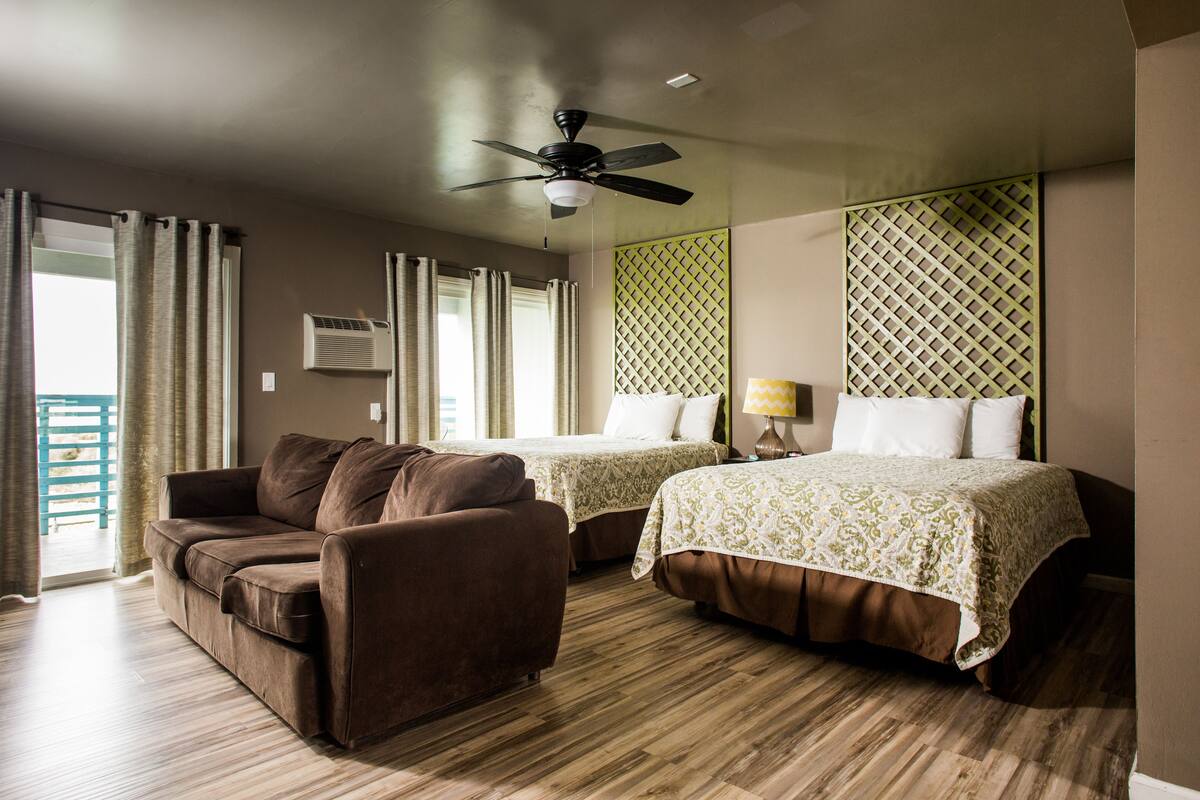 A hotel room with a brown sofa, two single beds with patterned bedspreads, a ceiling fan, and a view of the outdoors.