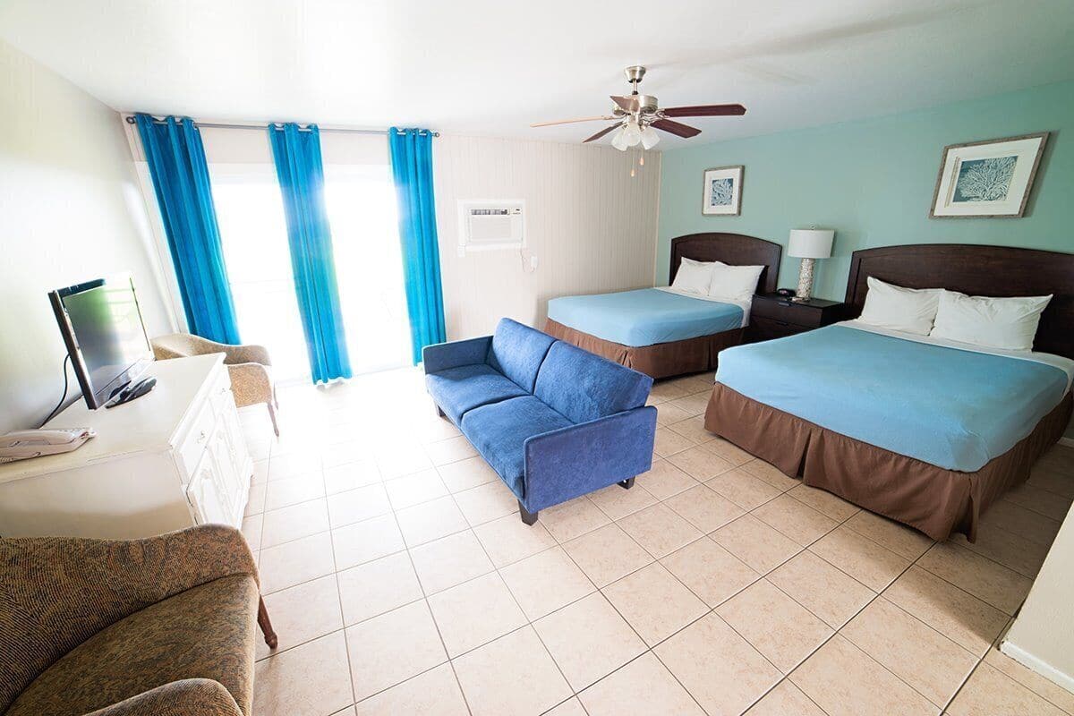 A hotel room with two beds, a sofa, a desk with a computer, and a ceiling fan.