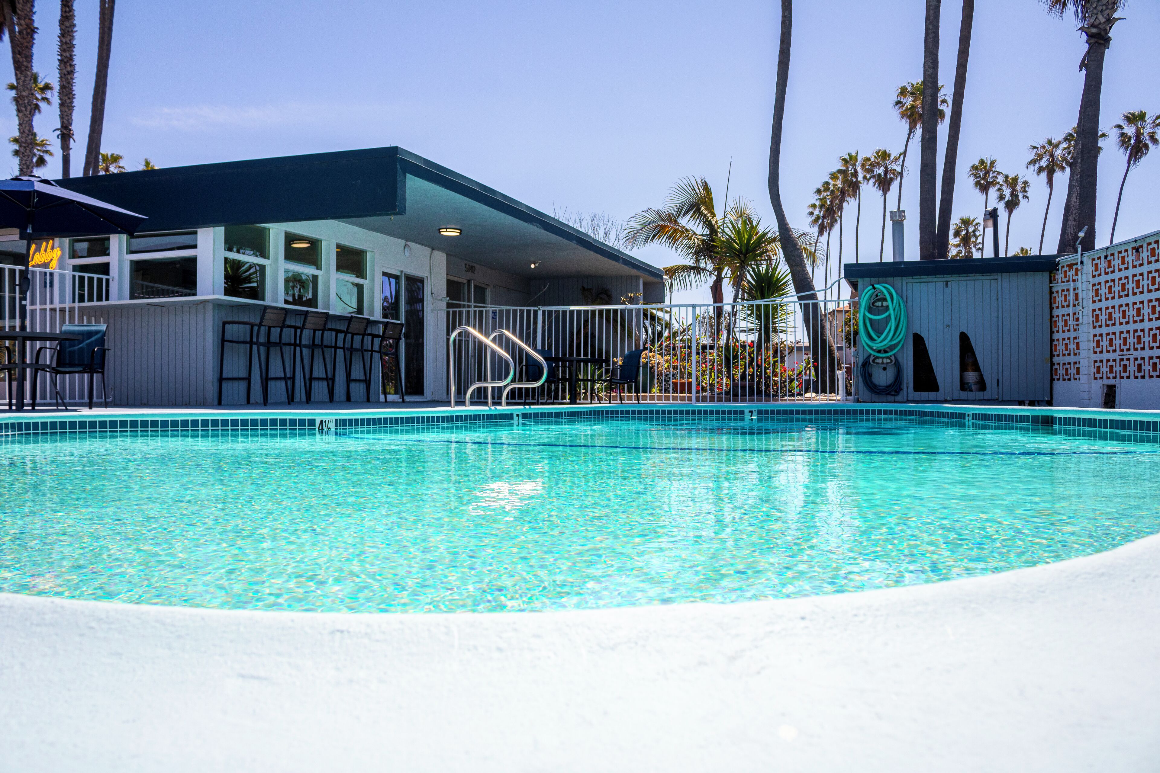 Piscina com área de bar, palmeiras e um edifício com letreiro neon.