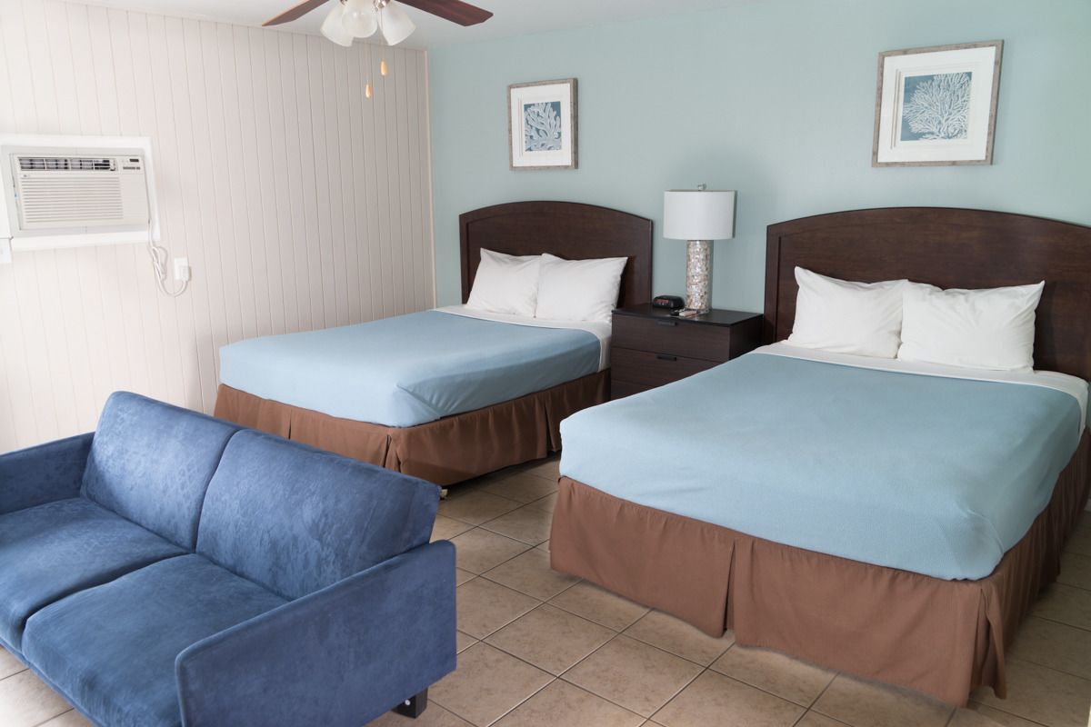 Een hotelkamer met twee bedden, een blauwe bank, een plafondventilator en kunstwerken in lijsten aan de muur.