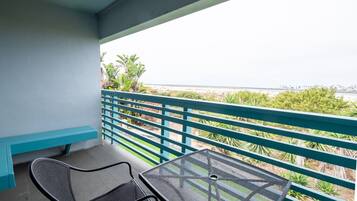 A balcony with a table and two chairs, overlooking a beach with greenery and a distant cityscape.