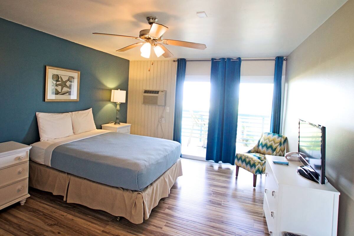 A hotel room with a bed, a desk, a chair, a ceiling fan, and a window with curtains.