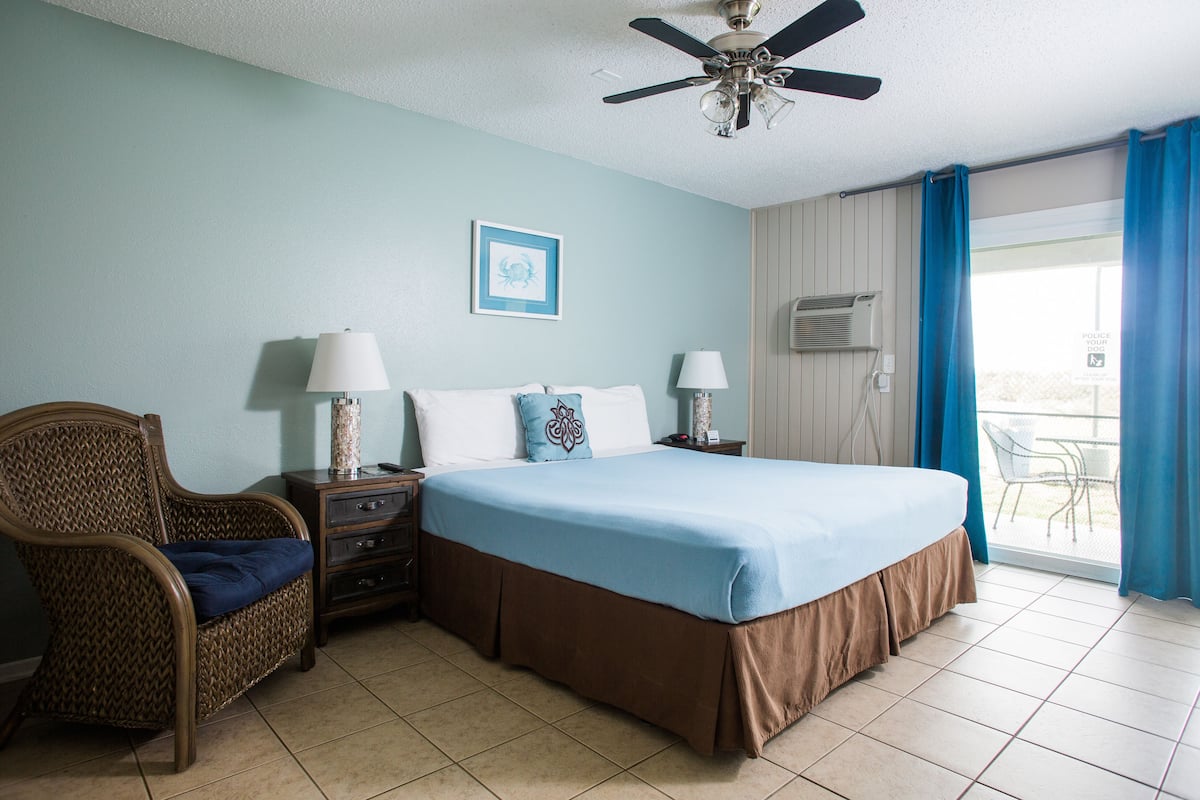 A hotel room with a bed, a chair, two bedside tables with lamps, a ceiling fan, a wall-mounted air conditioner, and a window with curtains.