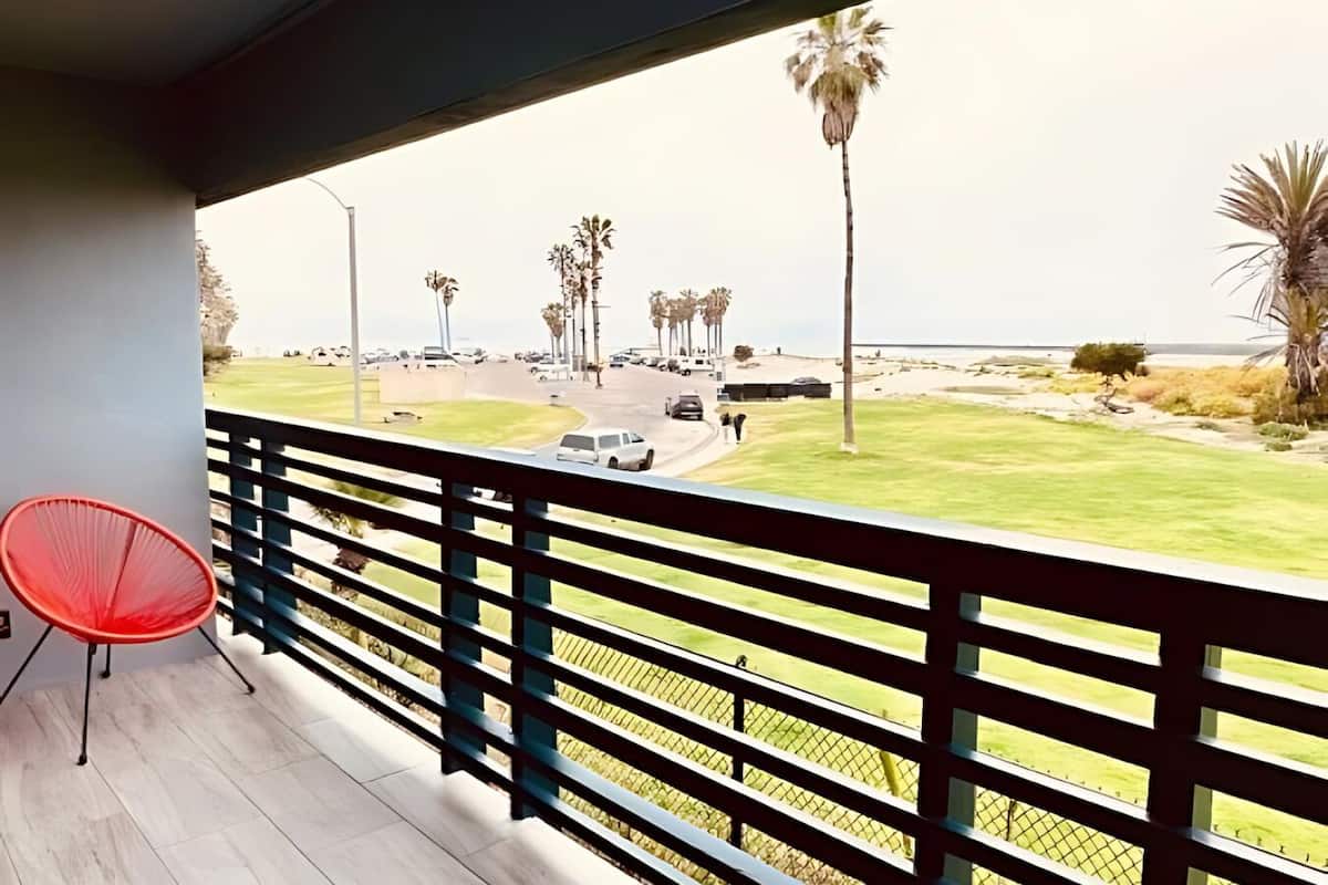 A balcony with a red chair, overlooking a road with palm trees and a grassy area.