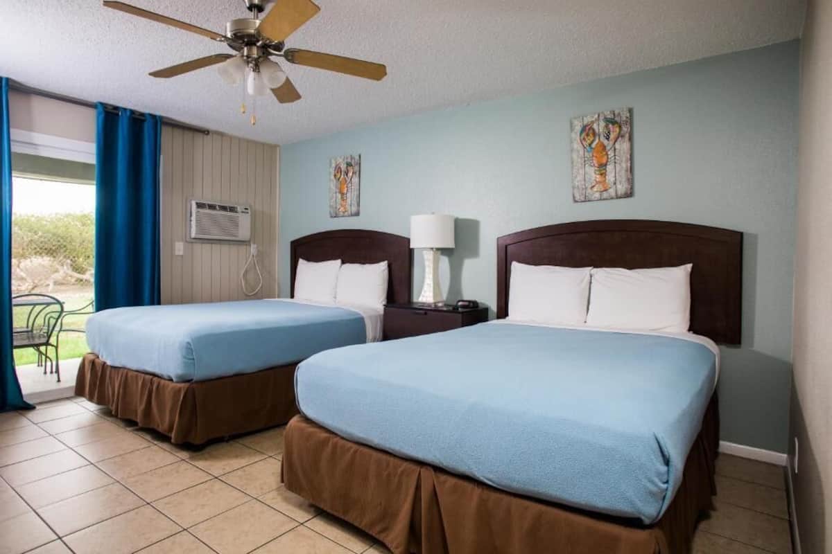 A hotel room with two beds, a ceiling fan, a window with curtains, and artwork on the walls.