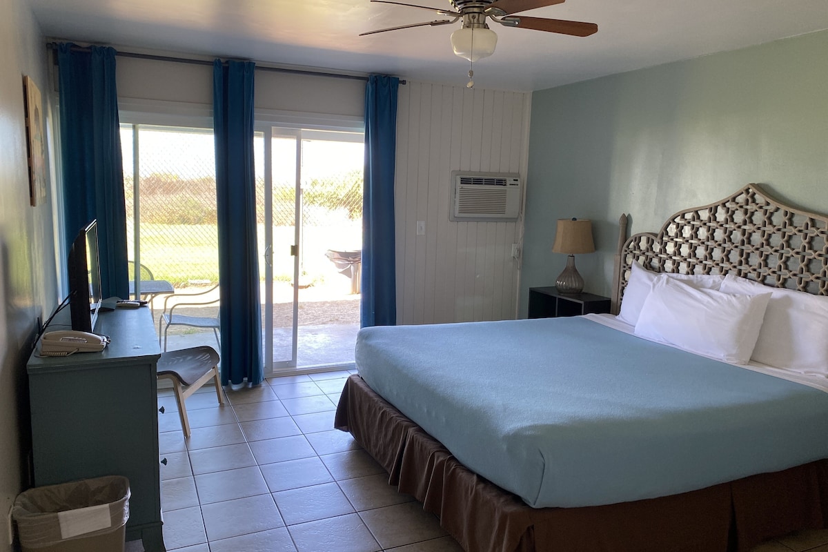 A hotel room with a bed, a desk with a lamp, a ceiling fan, and a view through a sliding door to an outdoor area.