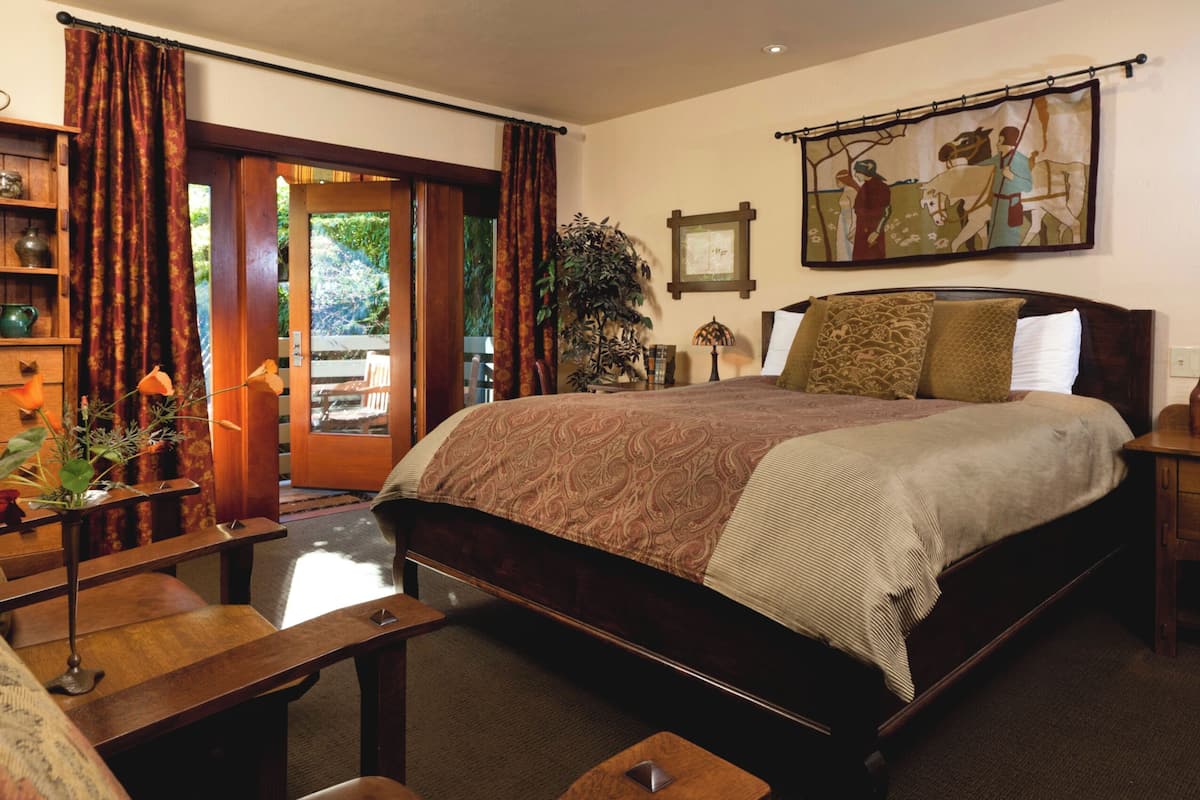 A bedroom with a large bed, wooden headboard, a nightstand, a desk, and a chair. There is a window with curtains, a plant, and a painting on the wall.