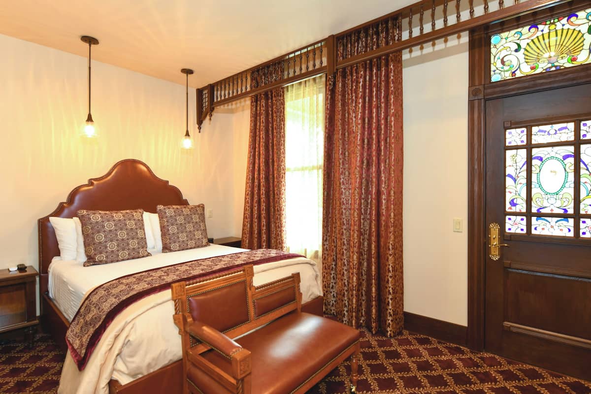 A bedroom with a large bed, a wooden bench, a nightstand, and a door with stained glass.