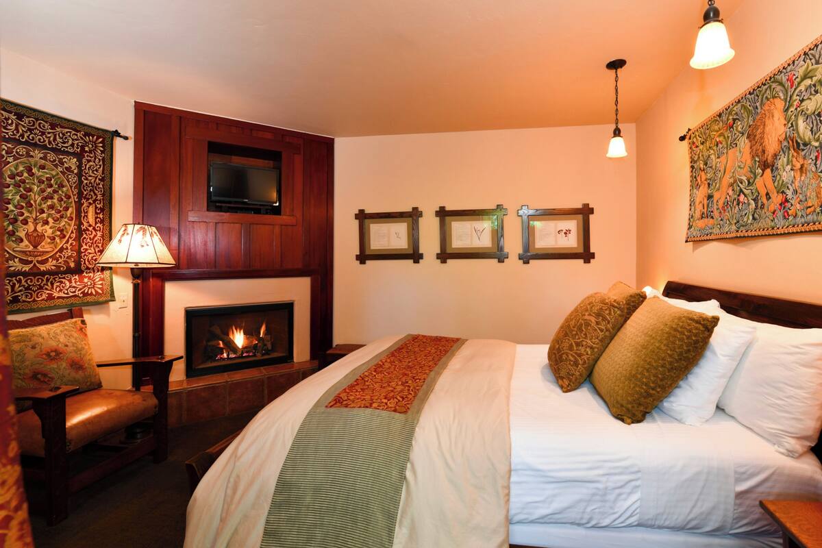 A cozy bedroom with a fireplace, a bed with multiple pillows, a chair, and framed pictures on the wall.