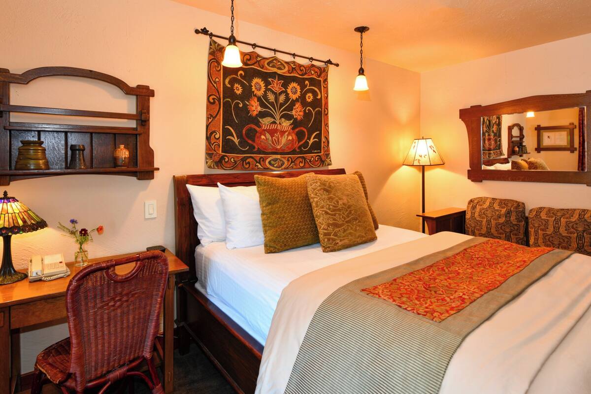 A hotel room with a bed, a desk with a lamp, a chair, a tapestry, and a mirror.