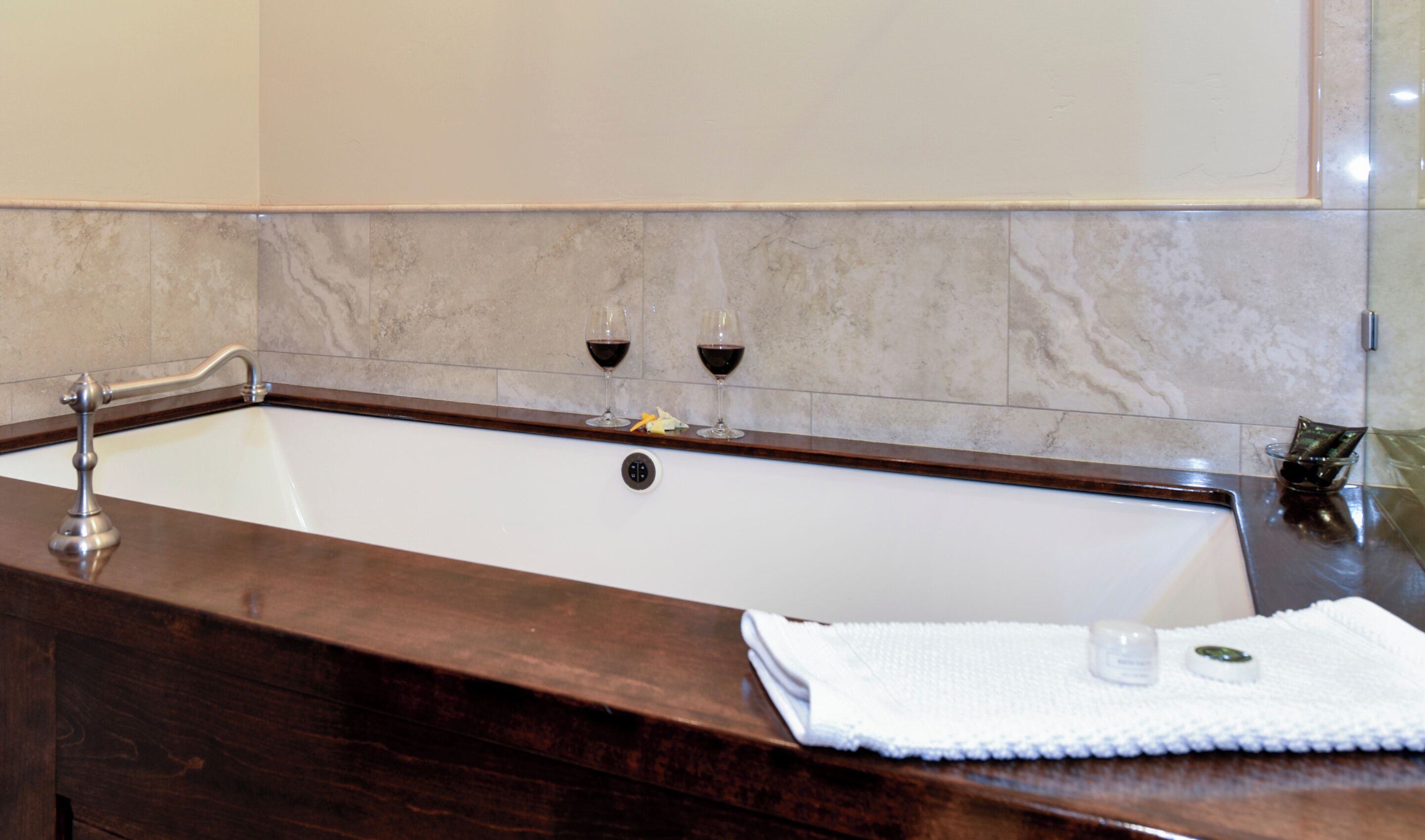 Une salle de bain avec un mur recouvert de carreaux de marbre, un meuble-lavabo en bois, une baignoire et deux verres à vin accrochés au mur.