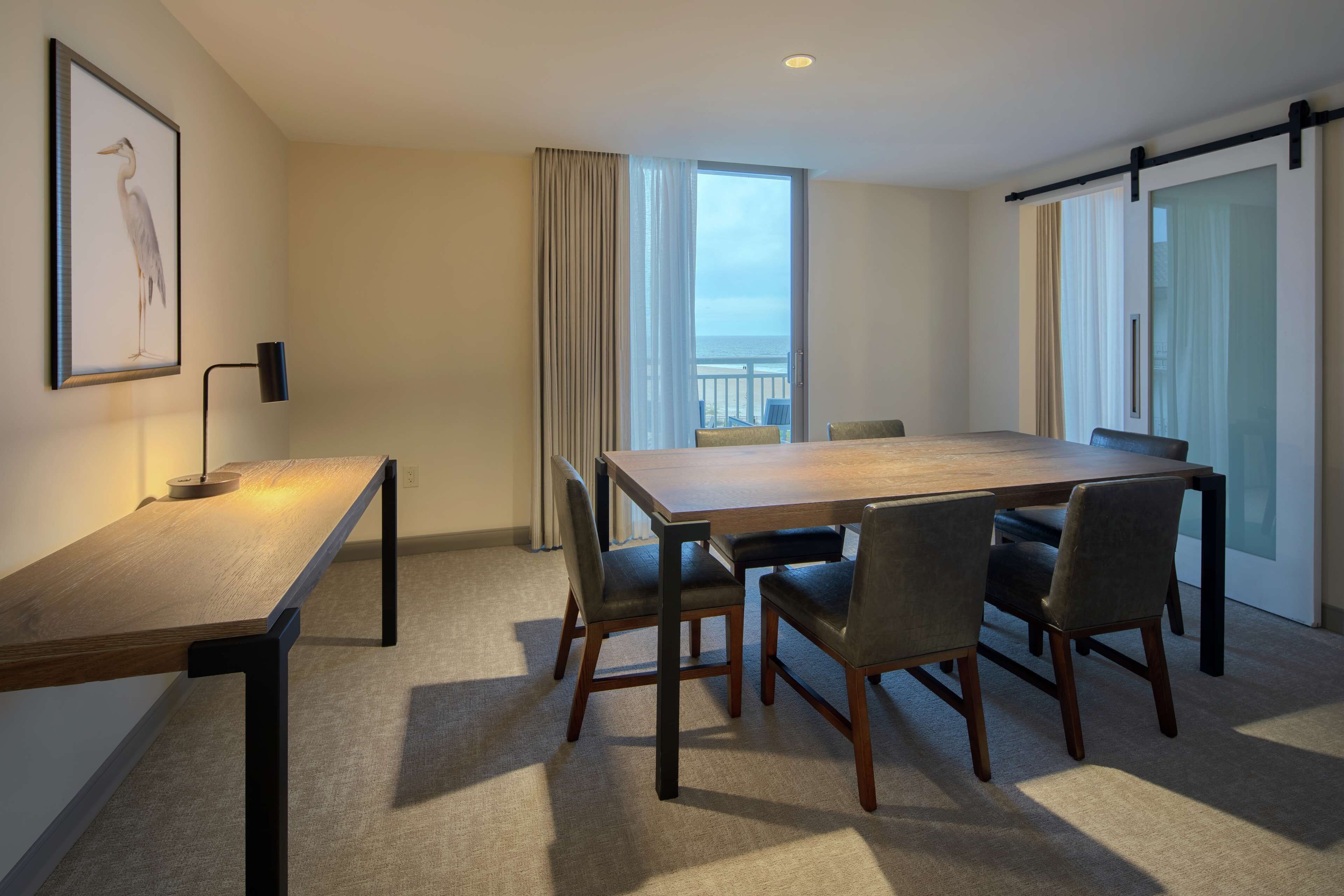 A hotel room with a wooden dining table, four chairs, a framed picture on the wall, a floor lamp, and a large window with curtains.