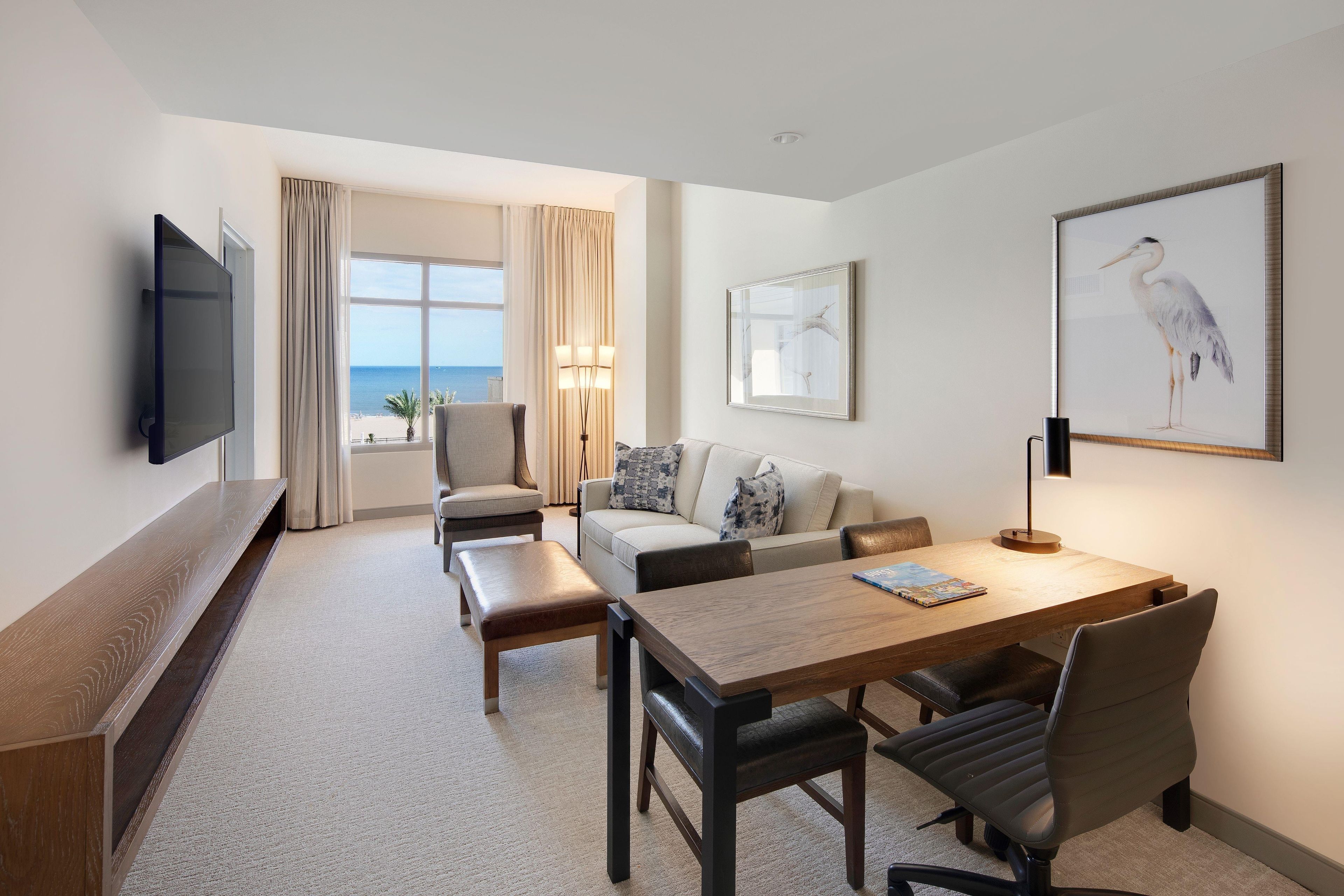 A hotel room with a flat-screen TV, a sofa, a dining table with chairs, a lamp, and a framed artwork.