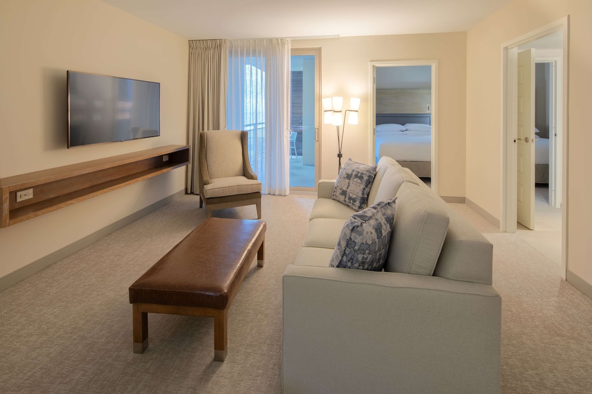 Une chambre d’hôtel équipée d’un téléviseur à écran plat, d’un banc en bois, d’un canapé avec des coussins à motifs, d’une chaise et d’un lit donnant sur l’extérieur.