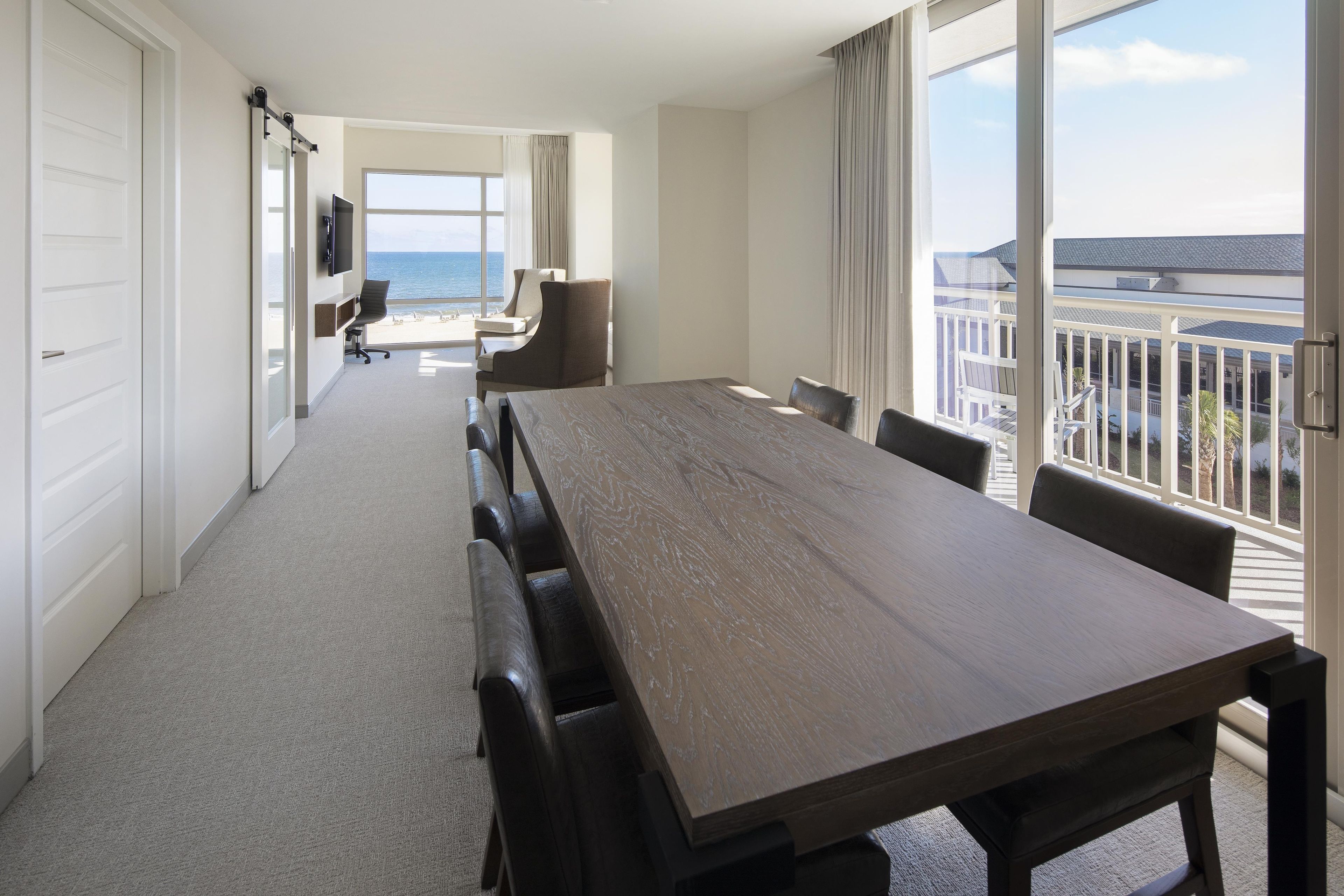 Habitación de hotel con una amplia mesa de comedor, un televisor montado en la pared y vista al océano a través de las ventanas.