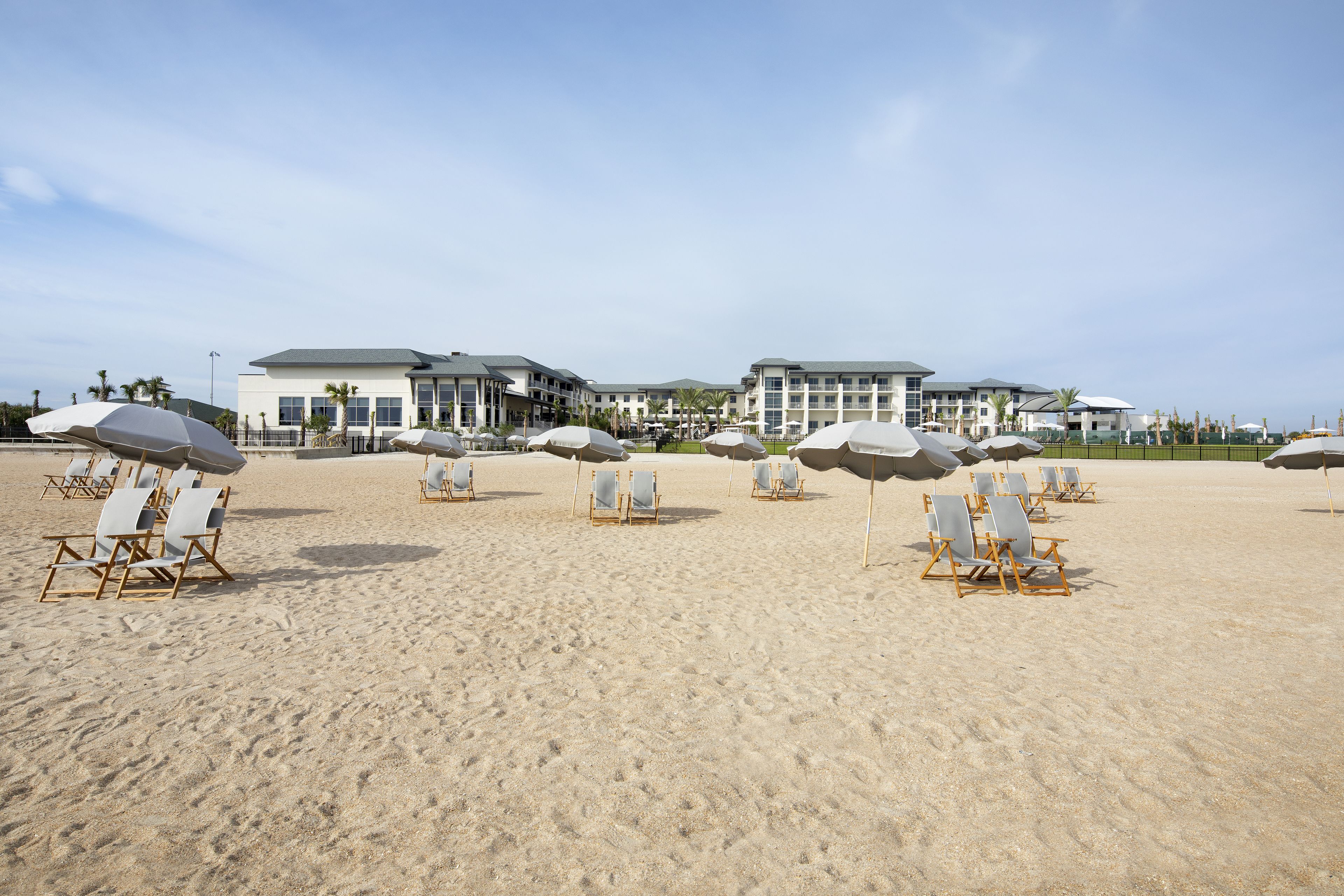 Une plage bordée de rangées de chaises de plage et de parasols, un grand bâtiment à étages et des palmiers en arrière-plan.