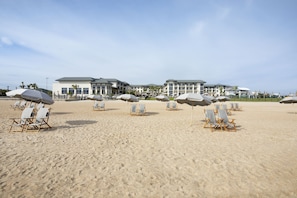 Une plage bordée de rangées de chaises de plage et de parasols, un grand bâtiment à étages et des palmiers en arrière-plan.