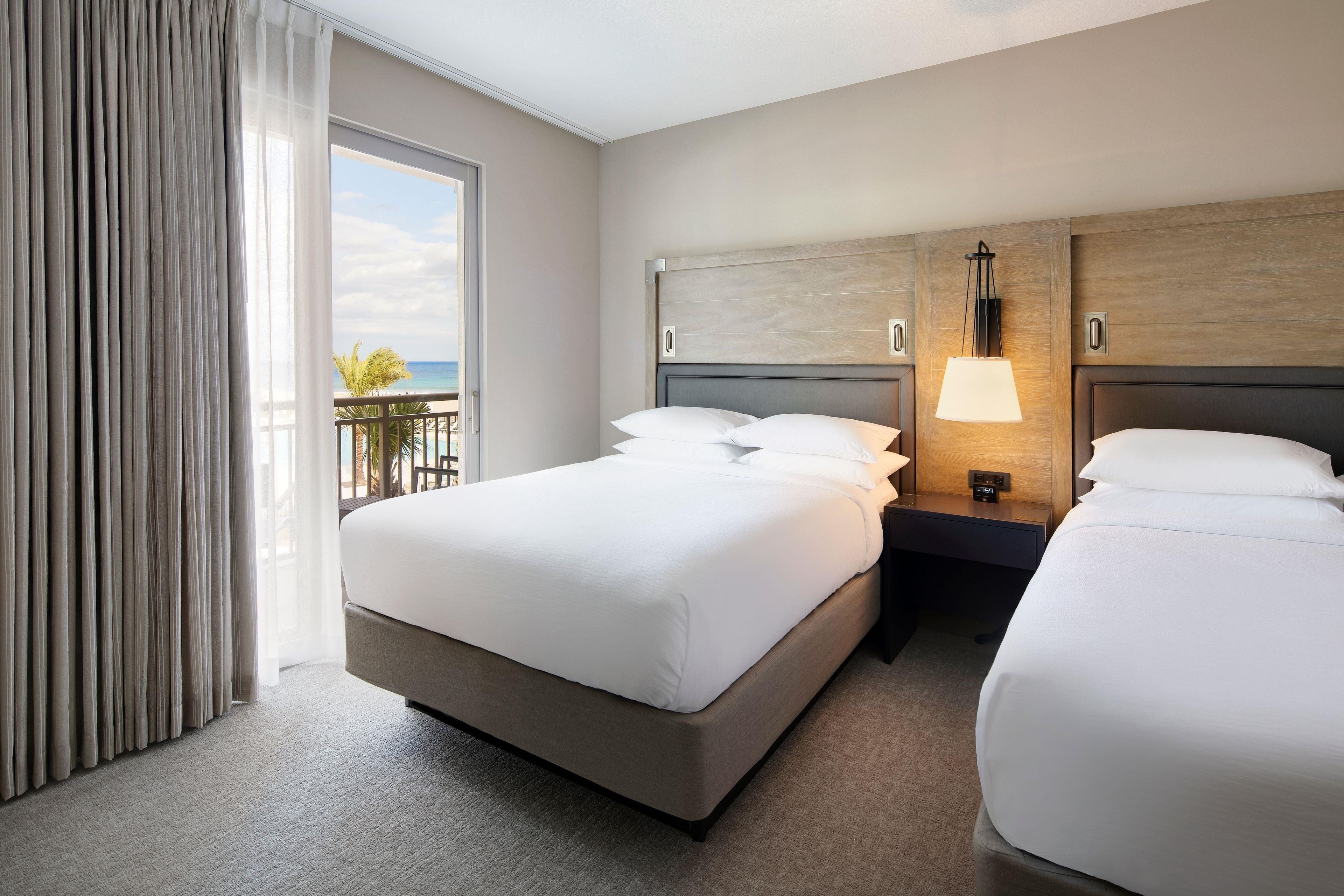 A hotel room with a large bed, a smaller bed, a nightstand with a lamp, and a view of the beach through the window.