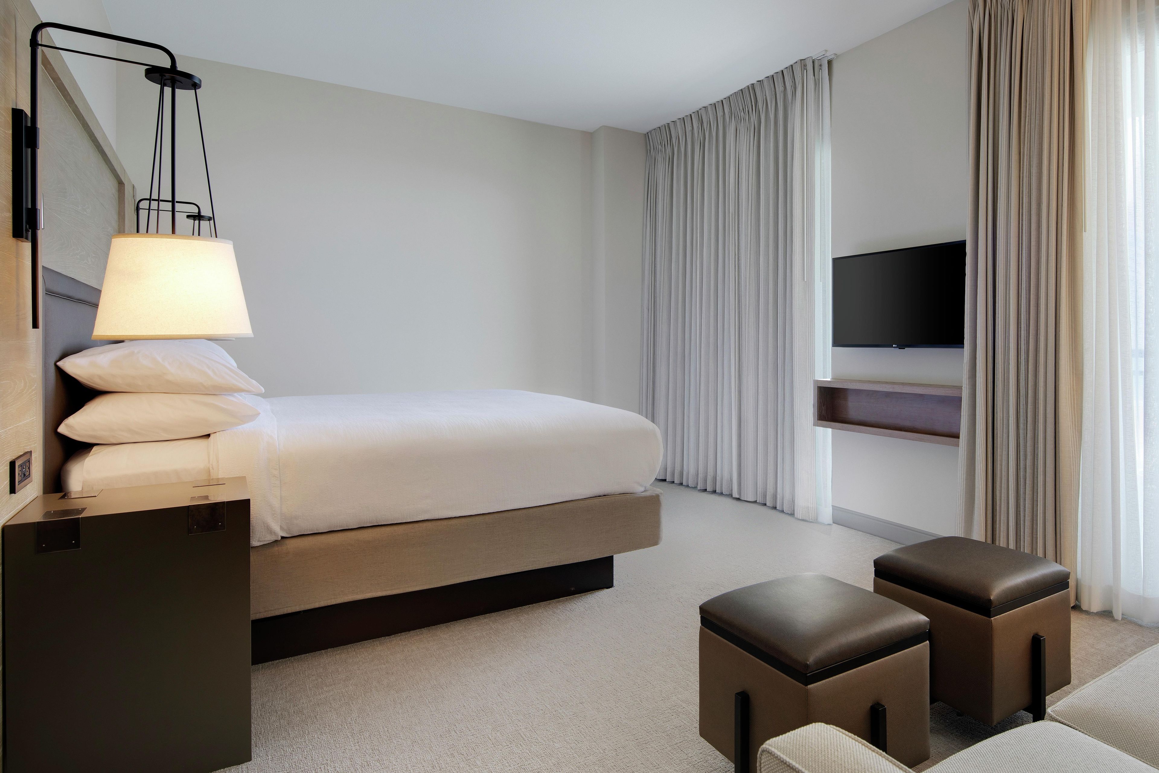 A hotel room with a bed, a bedside table, a lamp, a TV, and a seating area with a bench.