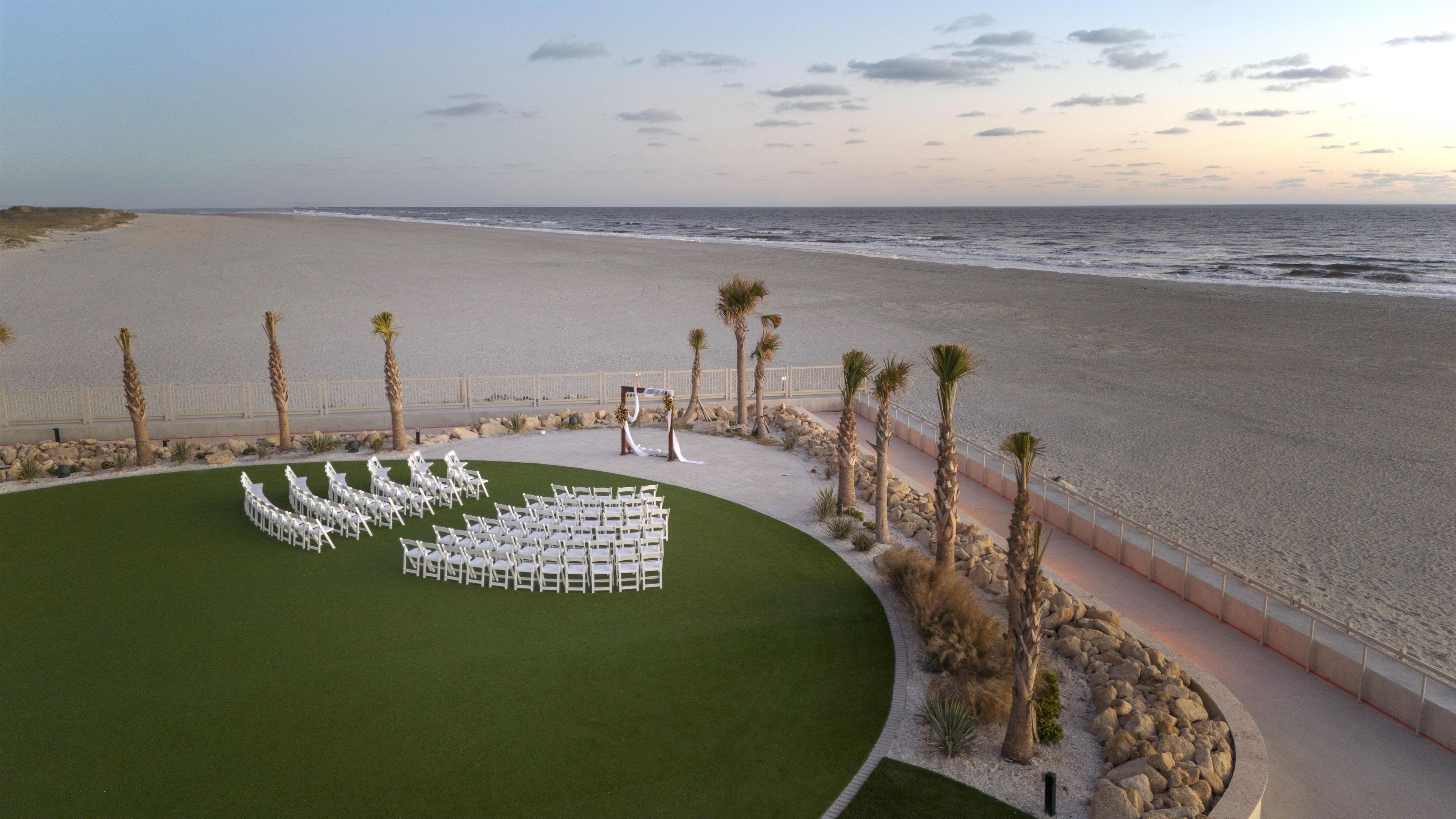 An outdoor wedding setup with chairs arranged in a semi-circle on a grassy area, a small altar, and palm trees along a beachfront.