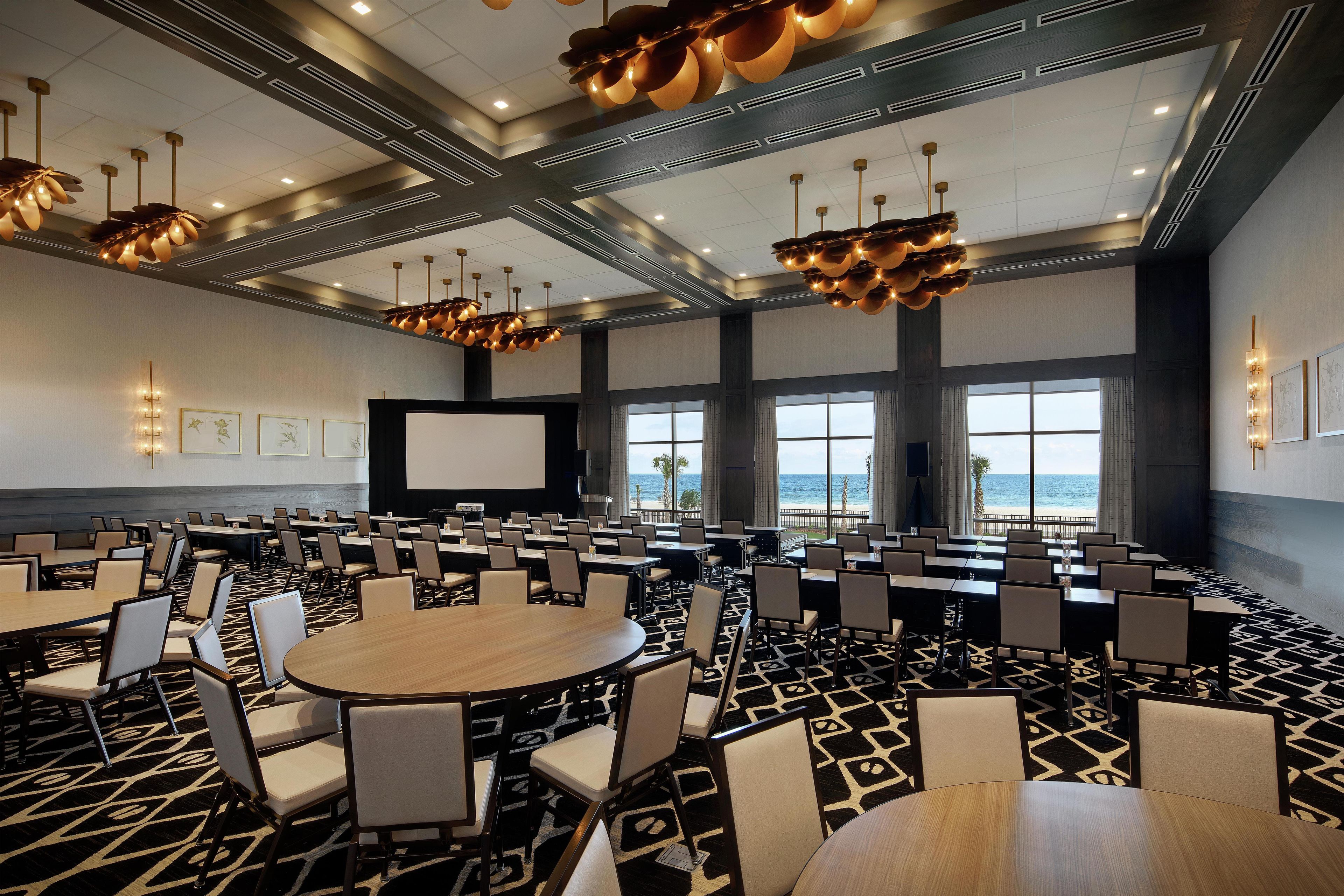 A conference room with multiple tables and chairs, a large screen, and a view of the ocean through large windows.
