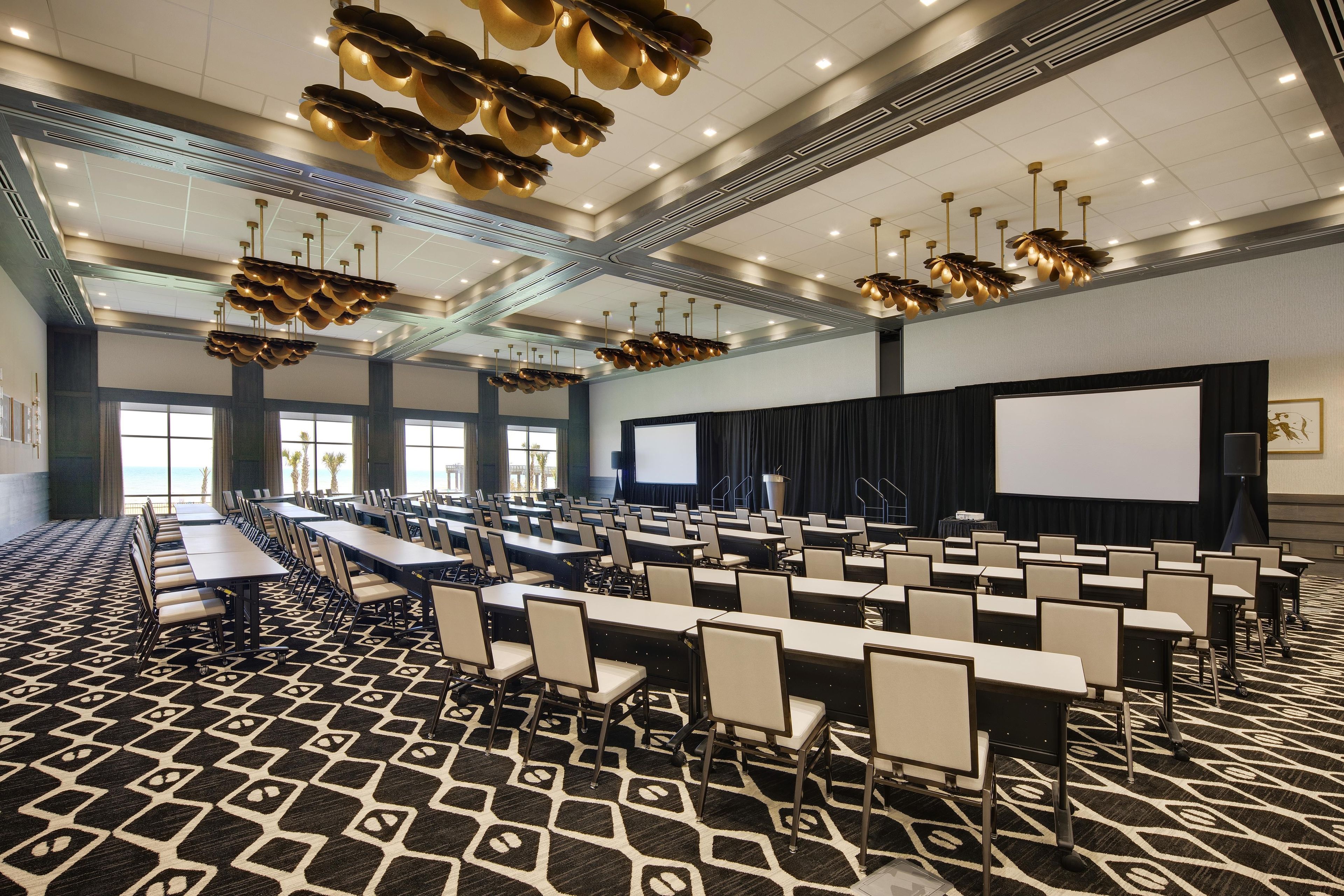 Une salle de conférence avec des rangées de tables et de chaises, de grandes fenêtres et un écran de projection.