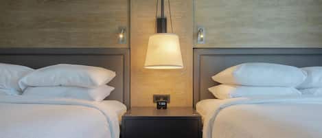 Quarto de hotel com duas camas, uma mesa de cabeceira com relógio e uma luminária na parede.