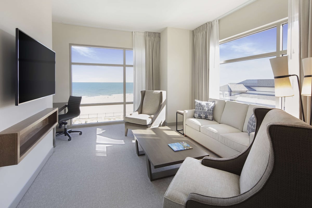 Une chambre d’hôtel dotée d’une grande fenêtre donnant sur la plage, d’un téléviseur à écran plat fixé au mur, d’un canapé, d’un fauteuil et d’une petite table d’appoint.