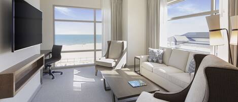 Habitación de hotel con un ventanal que ofrece vistas a la playa, un televisor de pantalla plana montado en la pared, un sofá, una silla y una mesita auxiliar.