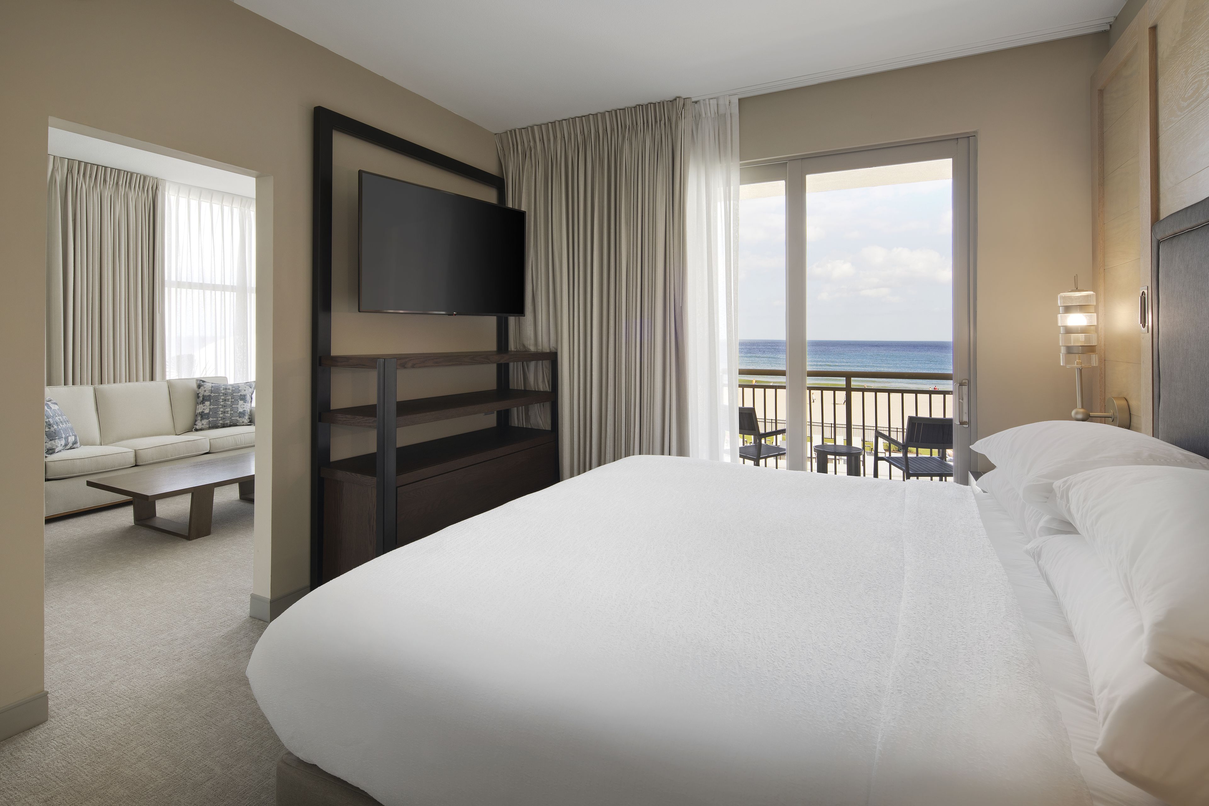 Una habitación de hotel con una cama grande, un televisor de pantalla plana en la pared, un balcón con vistas al océano y una zona de estar con sofá y sillones.