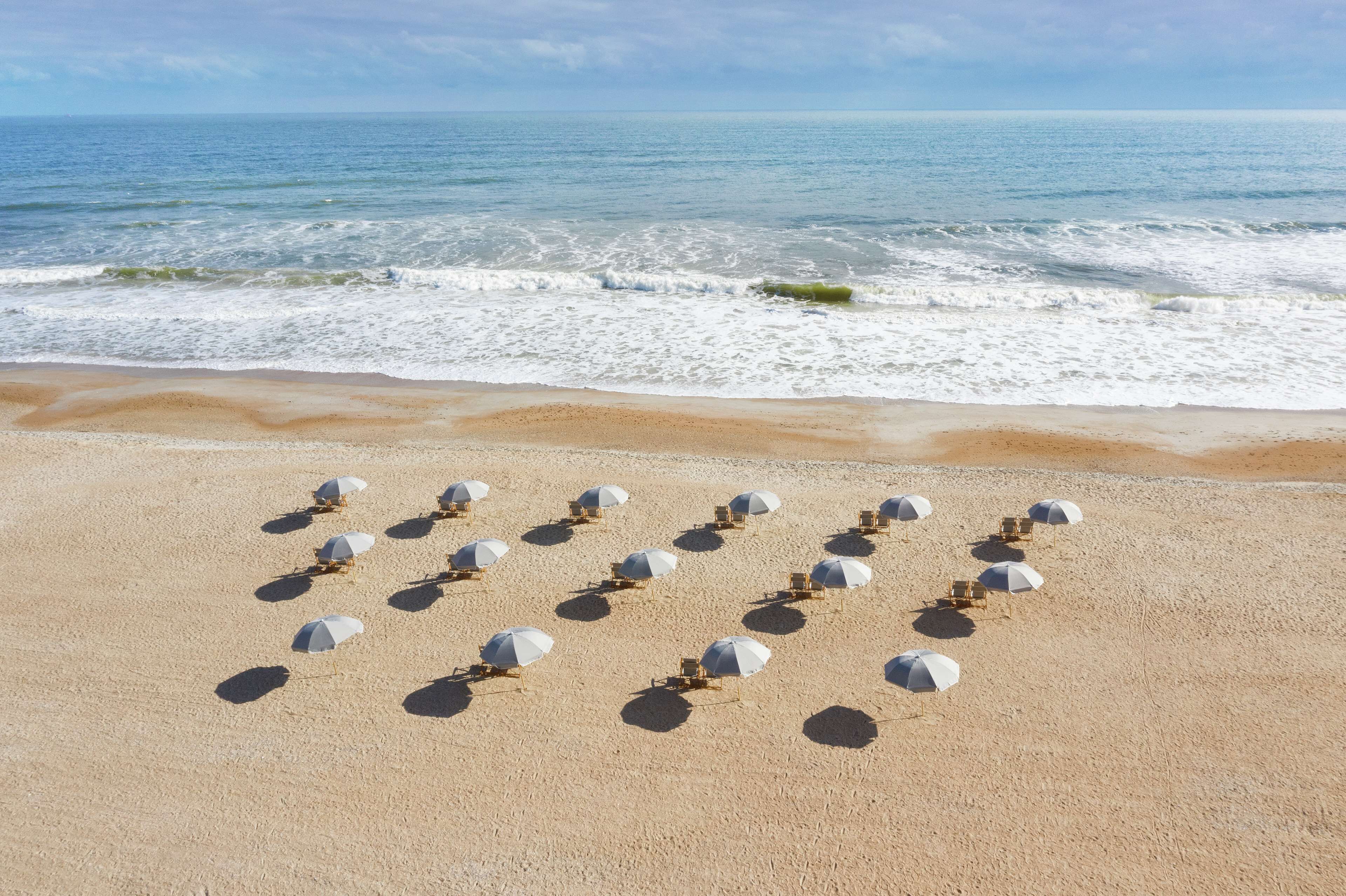 A beach with rows of sun umbrellas and lounge chairs aligned on the sand, facing the ocean.