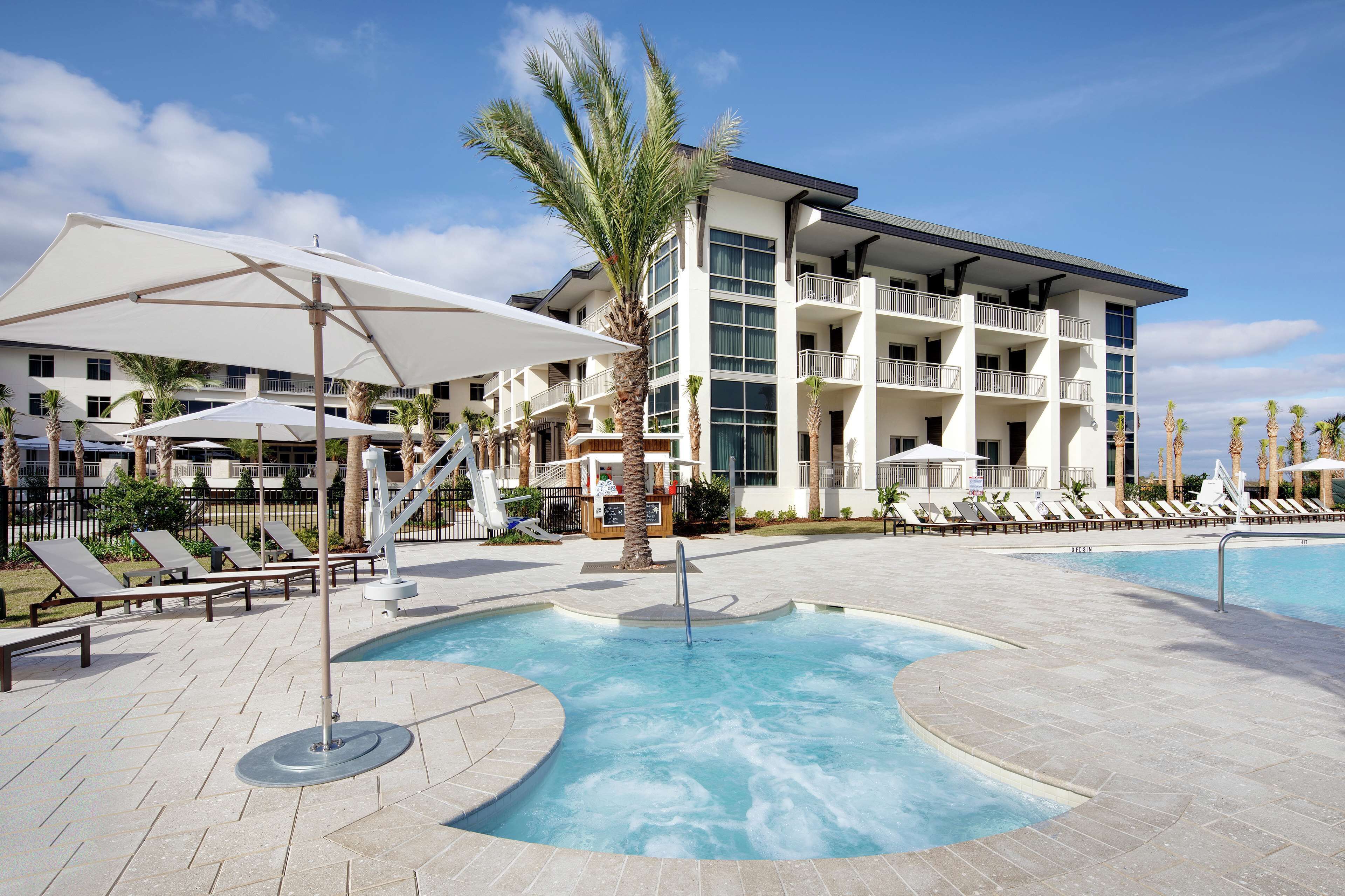 A pool area with lounge chairs and umbrellas, a palm tree, and a multi-story building with balconies.