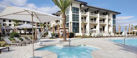 A pool area with lounge chairs and umbrellas, a palm tree, and a multi-story building with balconies.